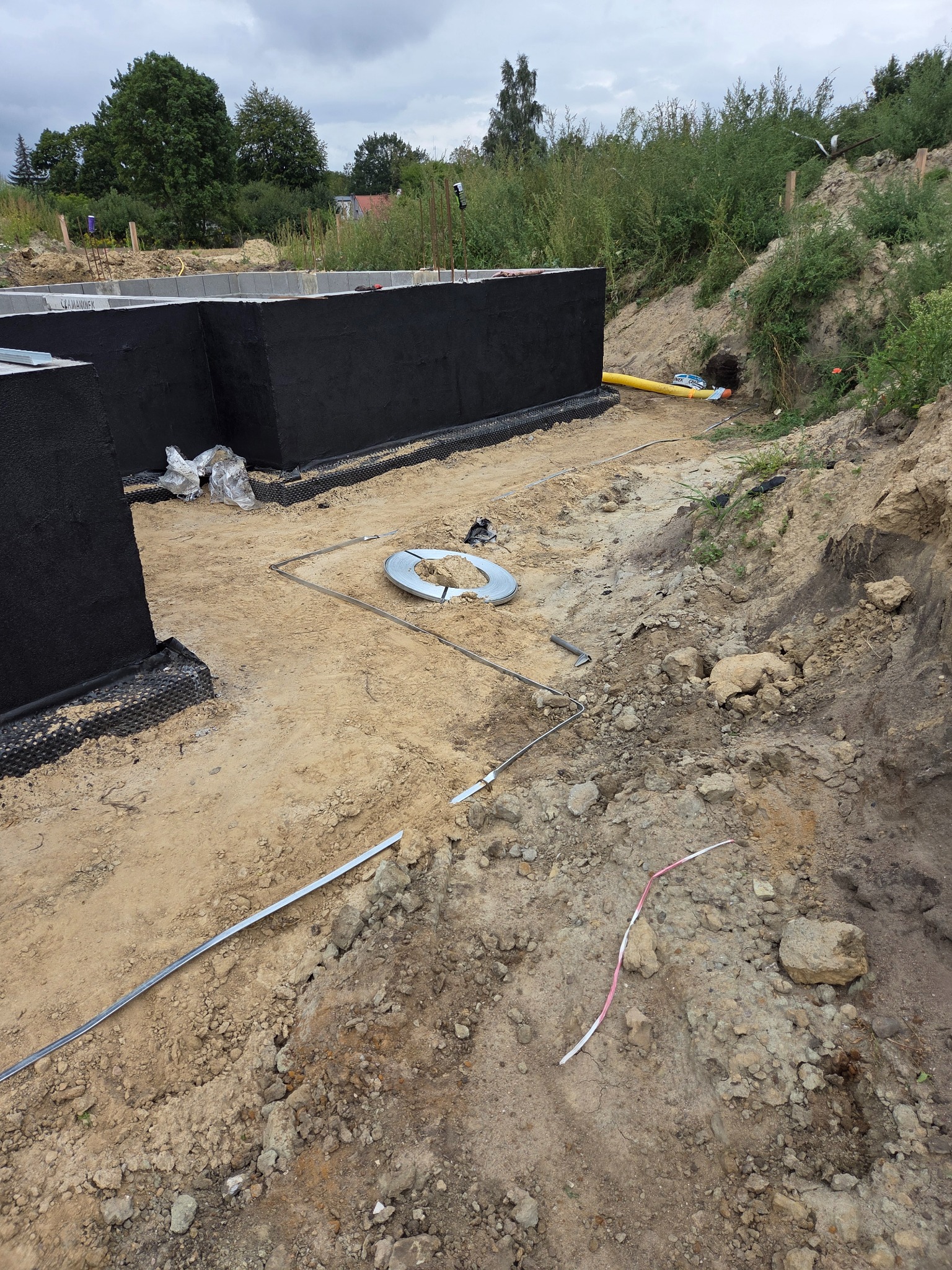 Fundamenty domu z izolacją przeciwwilgociową, widoczne rury i elementy instalacji w wykopie. Prace przygotowawcze do dalszych etapów budowy.