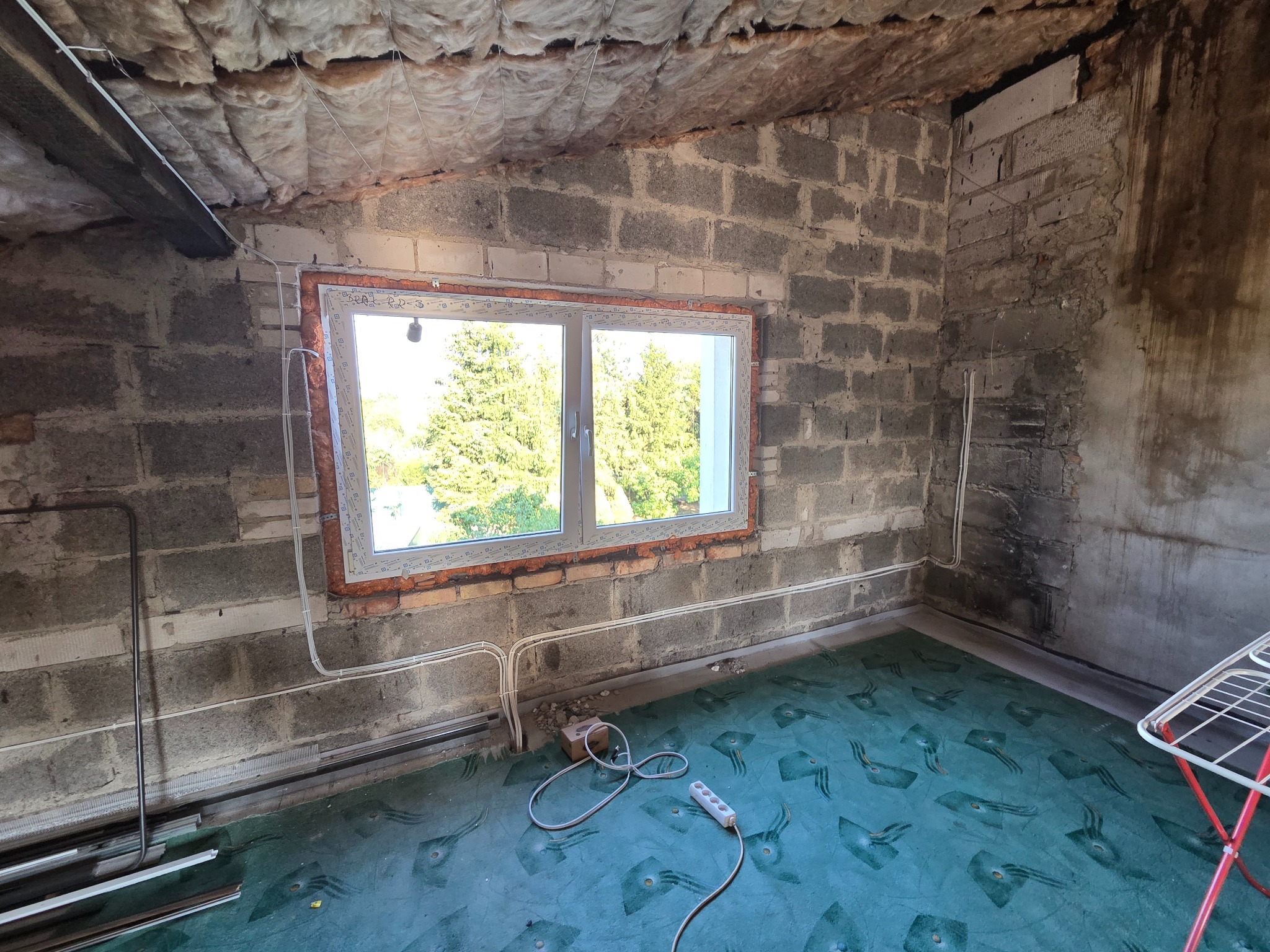 Surowy pokój na poddaszu z oknem, widoczne przewody elektryczne, ściany z bloczków, izolacja stropu. Gniazdko elektryczne na podłodze. Etap prac wykończeniowych.