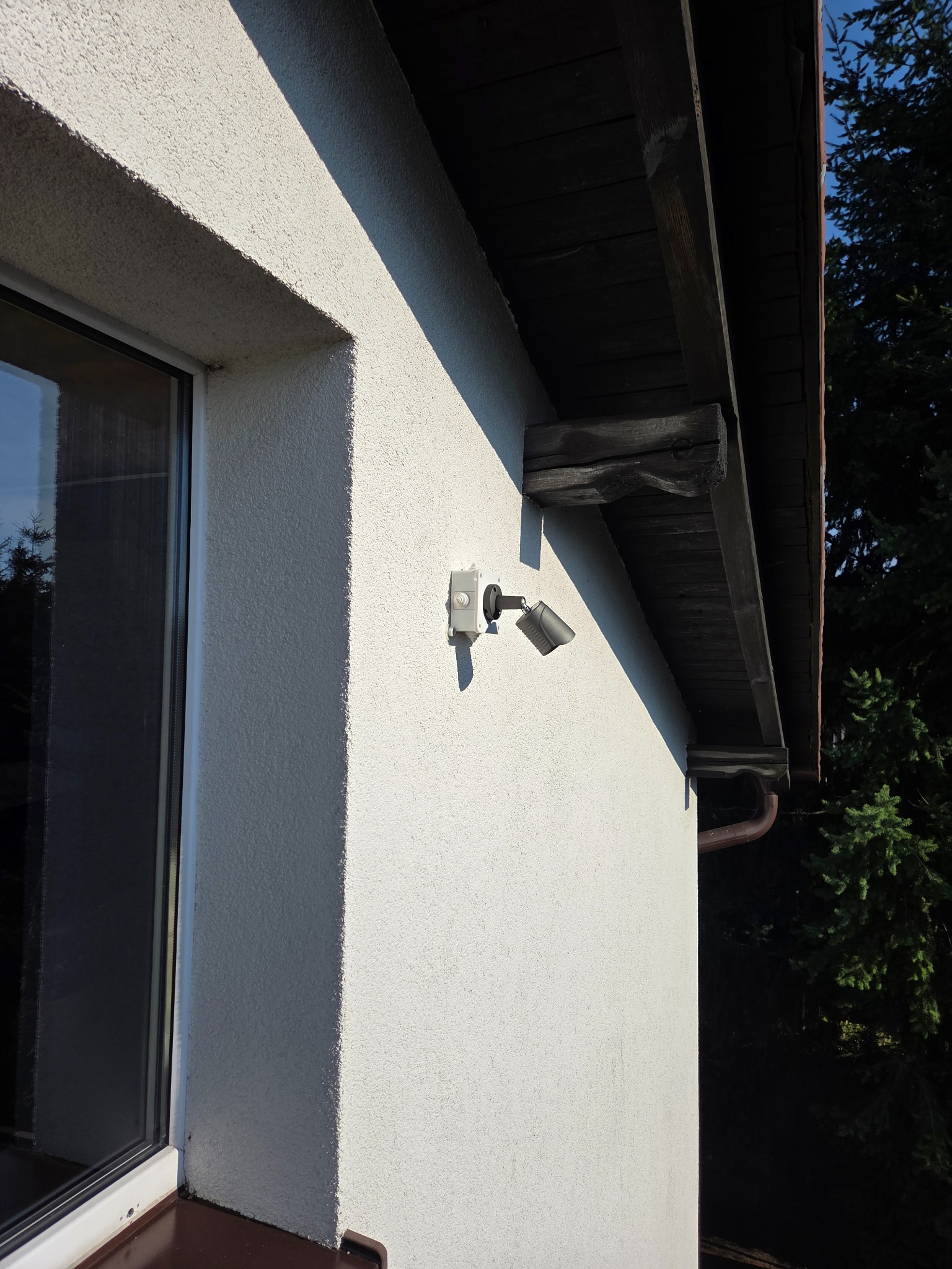 Zewnętrzny reflektor LED zamontowany na elewacji domu z oknem i drewnianym okapem dachu. Widoczny fragment rynny i zieleń w tle.