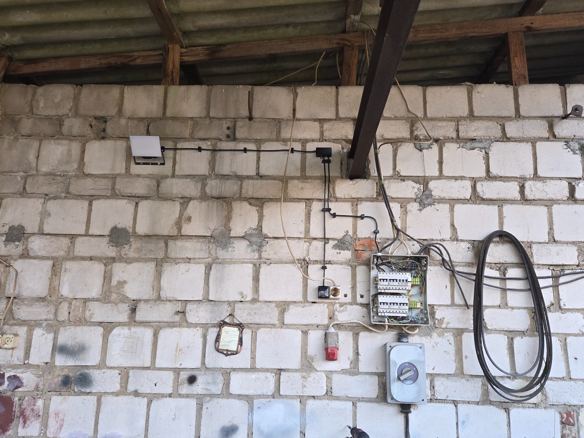 Otwarta skrzynka elektryczna z bezpiecznikami na białej ścianie z bloków, widoczne kable, gniazdka, włącznik i lampa. Elementy instalacji elektrycznej w surowym otoczeniu.