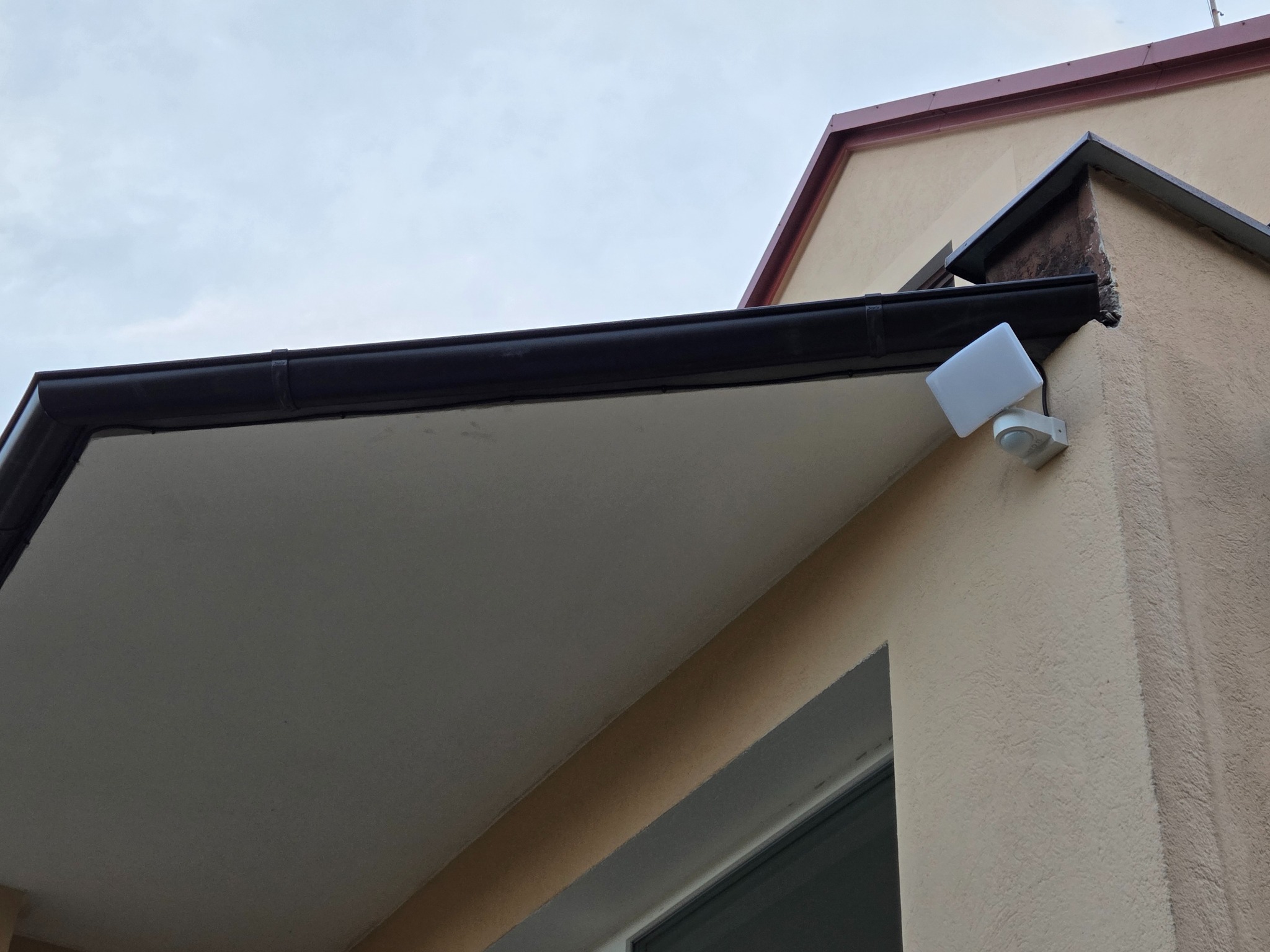 Zewnętrzny reflektor z czujnikiem ruchu zamontowany pod okapem budynku o beżowej elewacji. Widoczny fragment okna i rynna. Ujęcie z dołu, pochmurne niebo w tle.
