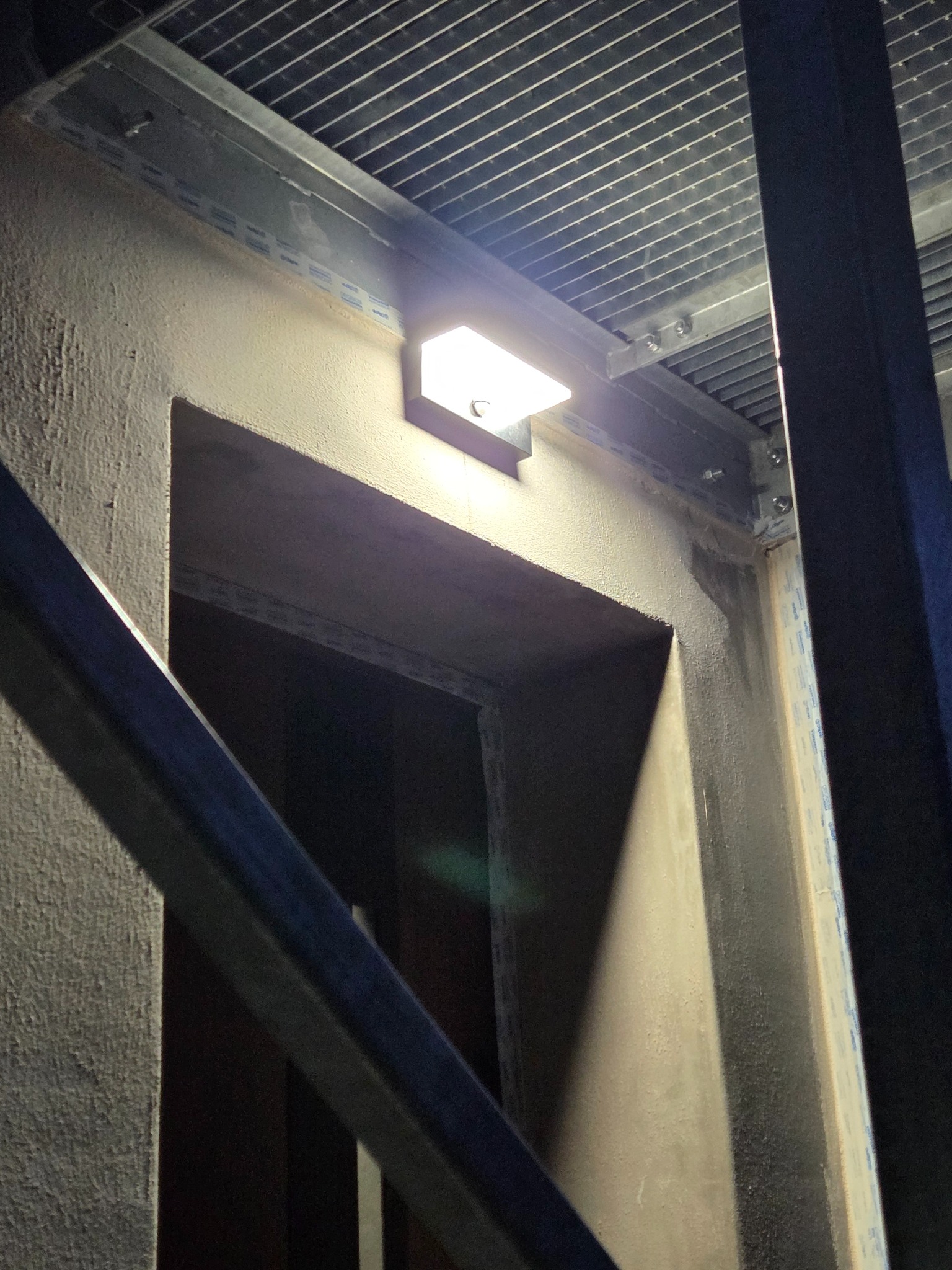 Zamontowana lampa zewnętrzna na szorstkiej ścianie obok otworu drzwiowego, z widoczną kratką sufitową i elementami konstrukcyjnymi, oświetlona od góry.