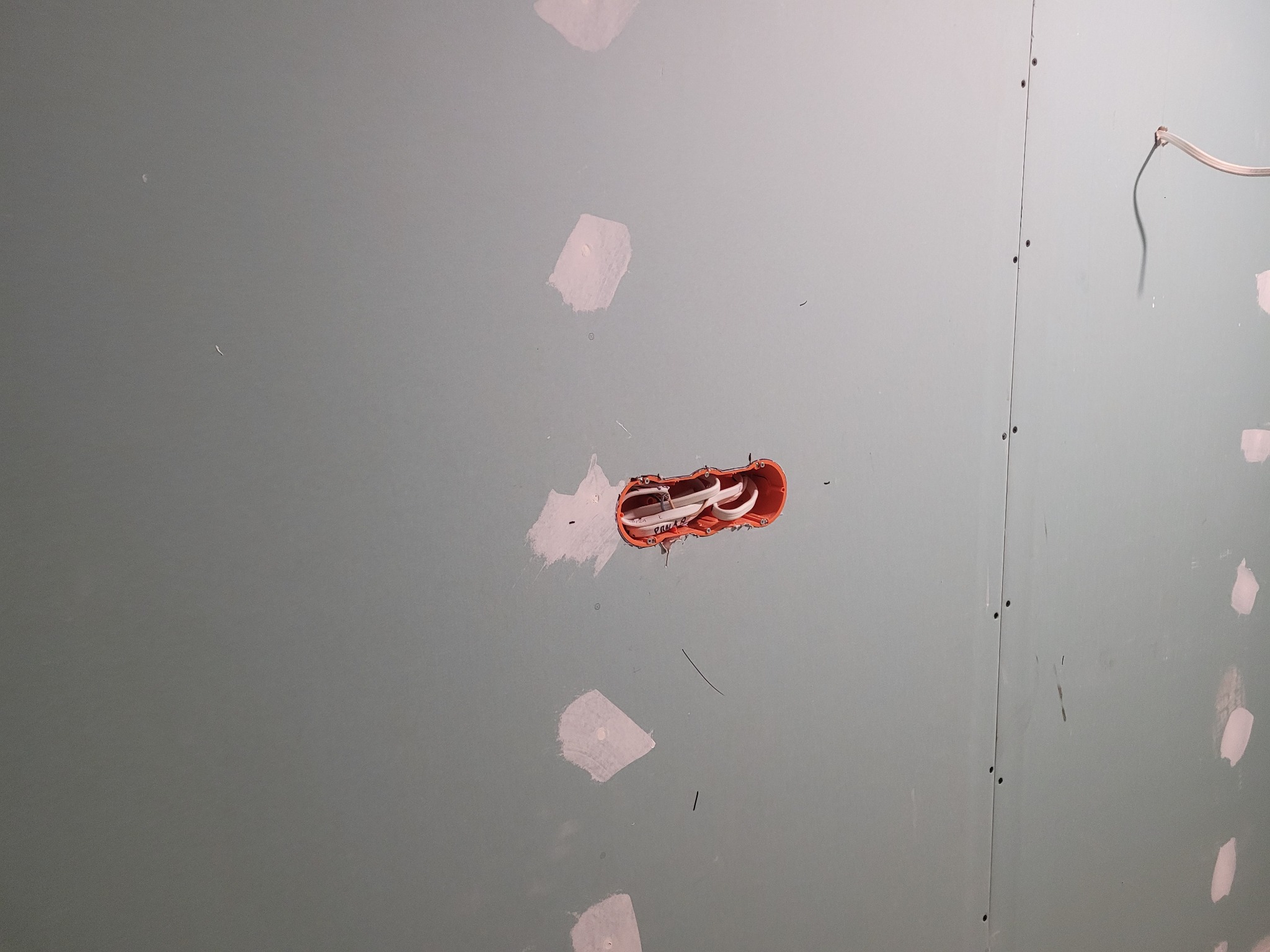 Ściana z płyt gipsowo-kartonowych z zamontowaną puszką elektryczną i przewodami, przygotowana do szpachlowania. Widoczne ślady po wkrętach i miejsca po szpachlowaniu.