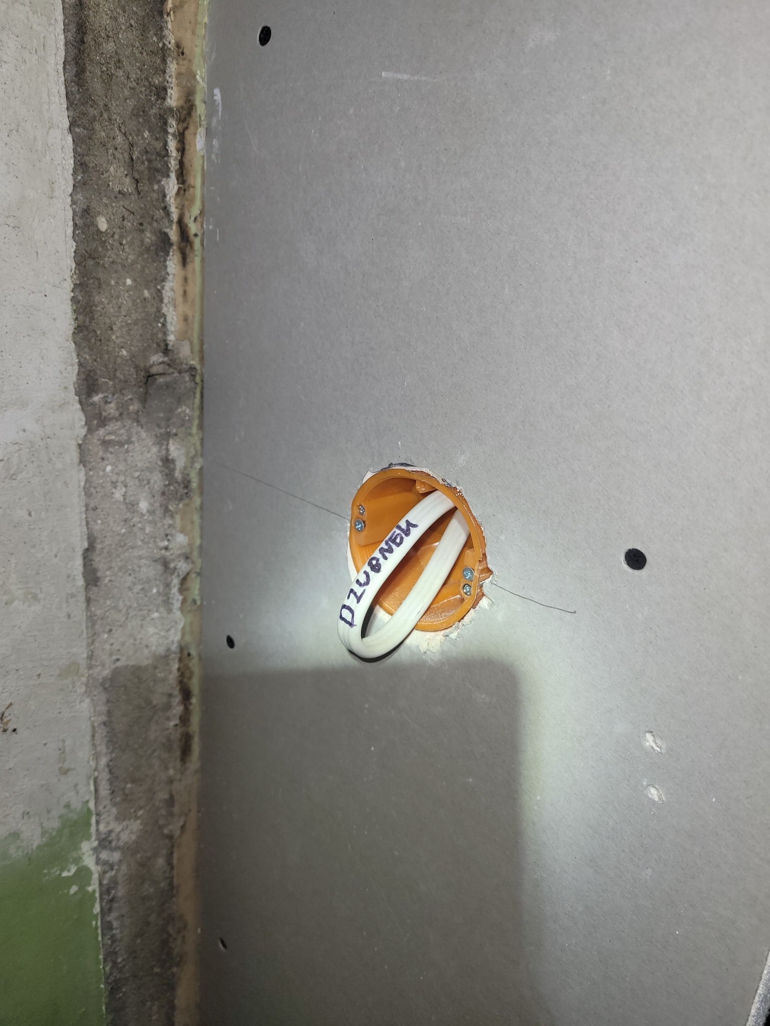 Pomarańczowa puszka elektryczna z kablami w szarej płycie gipsowej, obok surowy fragment ściany. Na kablu napis 'Dzwonek'.