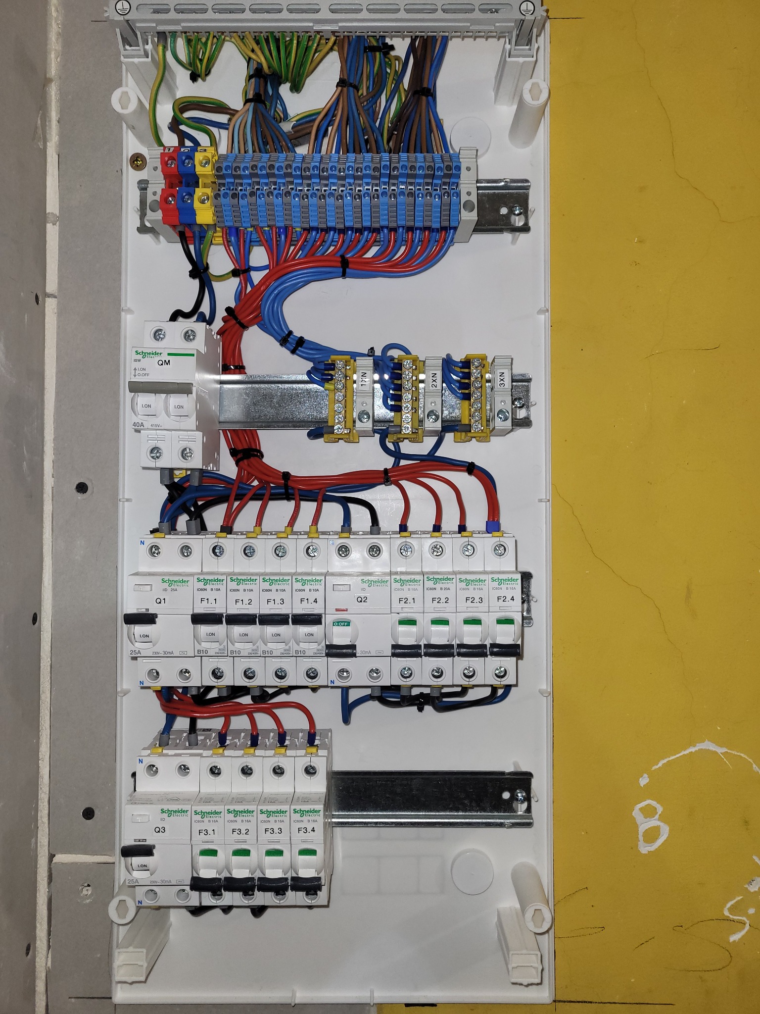 Rozdzielnica elektryczna w białej obudowie z widocznymi przewodami, bezpiecznikami i złączkami, zamontowana na ścianie. Widoczne elementy marki Schneider Electric.