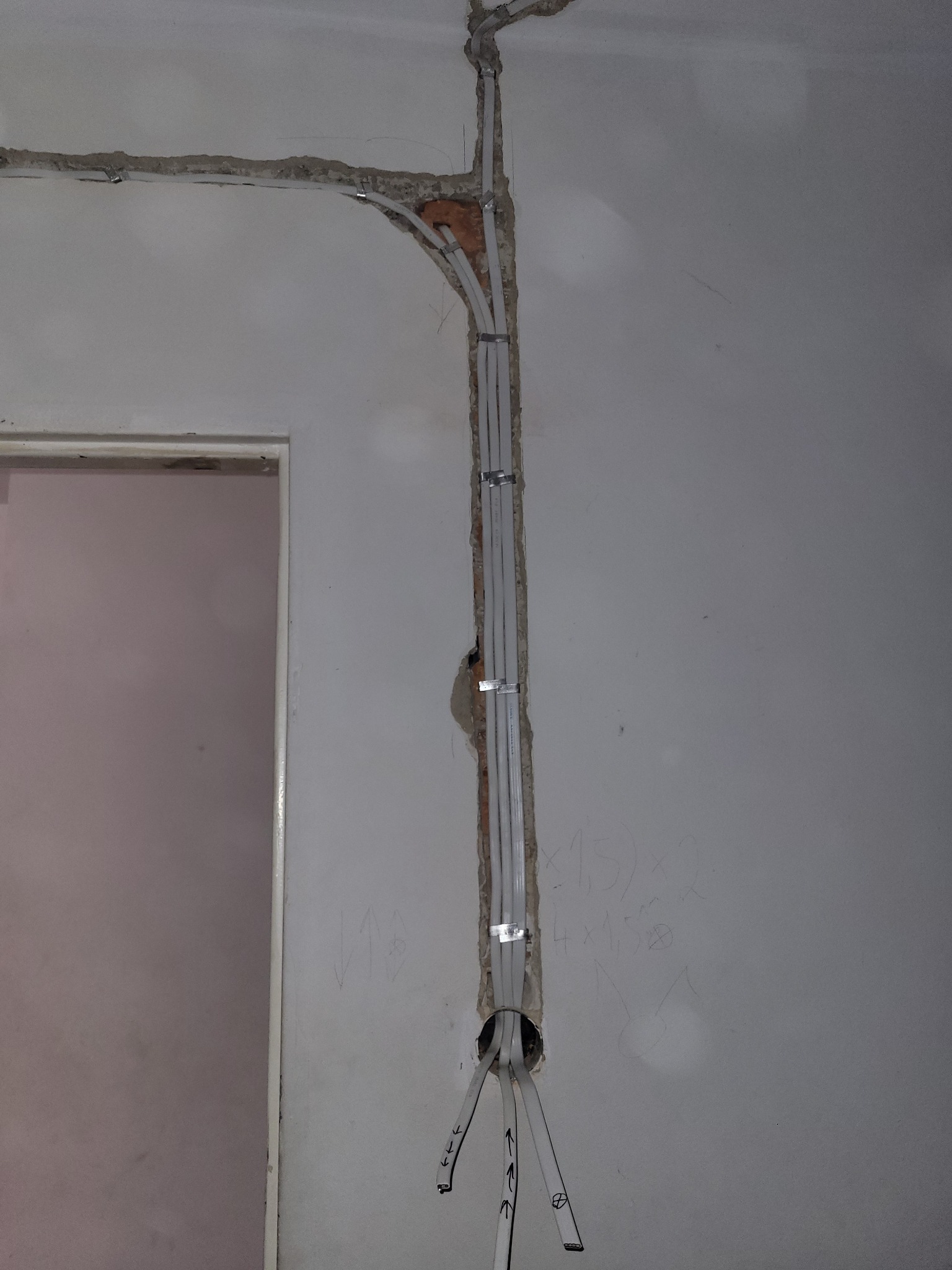 Białe kable elektryczne prowadzone w bruździe ściany, zabezpieczone uchwytami. Widoczne ślady po kuciu i rysunki na ścianie. Drzwi w tle po lewej.