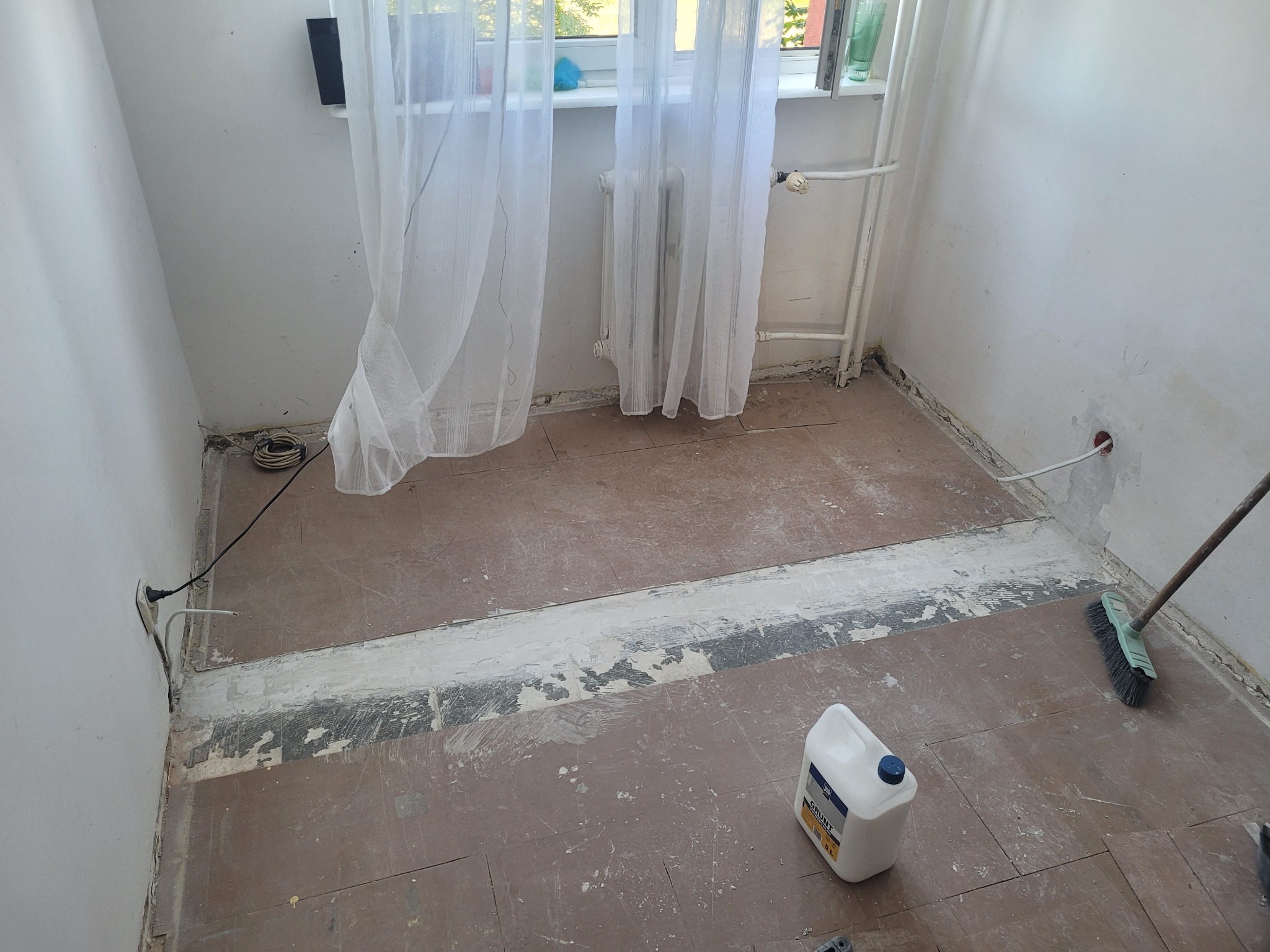 Przygotowanie podłoża: gruntowanie podłogi w pokoju z grzejnikiem i oknem. Widoczny kanister z gruntem i szczotka. Fragmenty starej podłogi i ściany w trakcie prac.