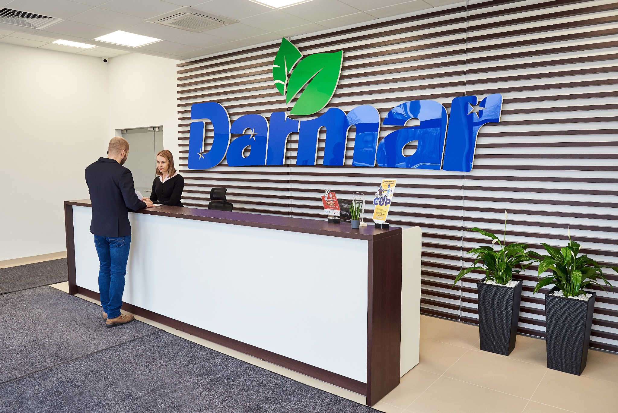 Recepcja biurowa z klientem i recepcjonistką. W tle logo firmy na ścianie z poziomymi panelami. Dwie rośliny doniczkowe. Czyste, nowoczesne wnętrze.