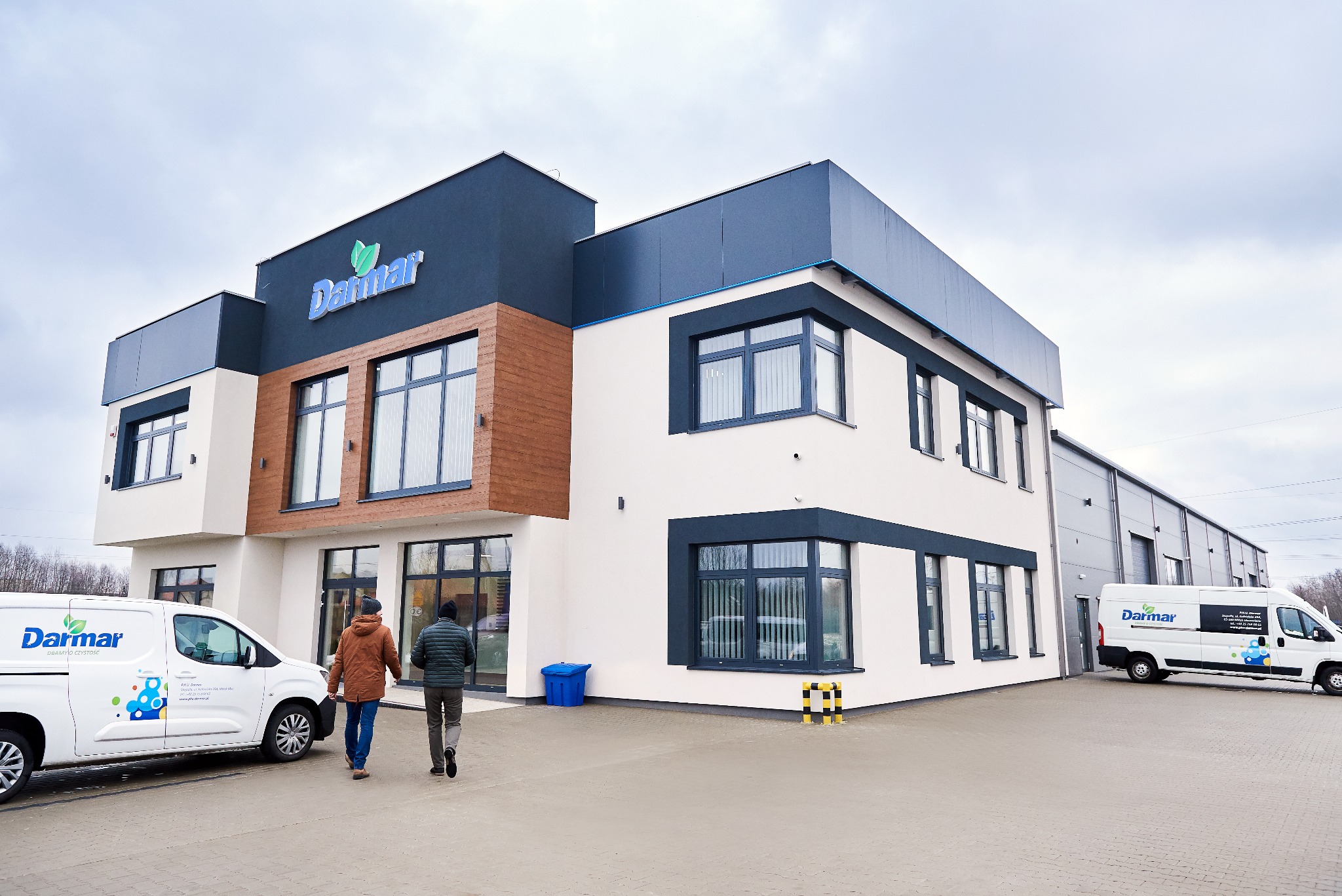 Budynek firmy Darmar z logo i dwoma busami firmowymi na zewnątrz. Dwie osoby idą w kierunku wejścia. Nowoczesna architektura, firmowe oznakowanie.