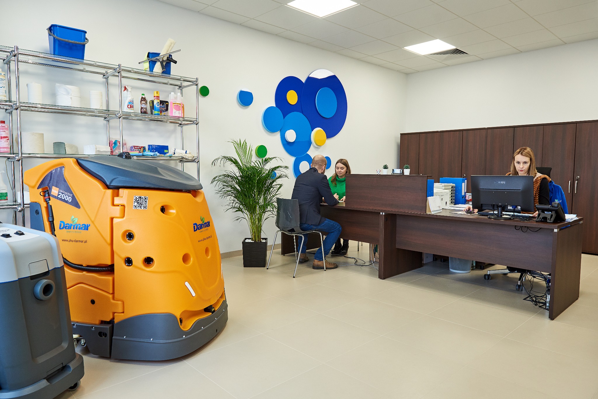 Biuro z pomarańczową maszyną czyszczącą Darmar Swingobot 2000, regałem z chemią i personelem przy biurku. Nowoczesne, jasne wnętrze z dekoracjami ściennymi.