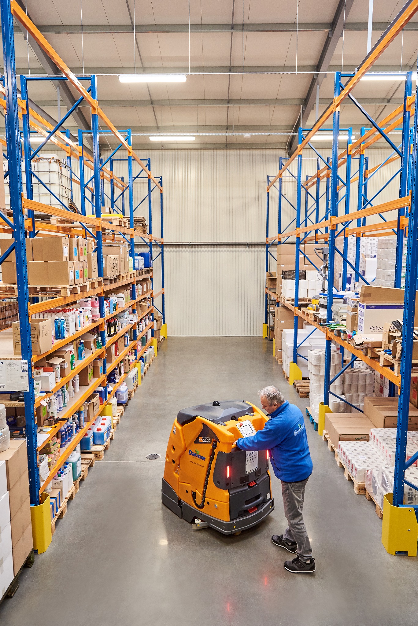 Pracownik obsługuje maszynę czyszczącą Darman WARRK 2000 w magazynie z regałami wypełnionymi towarem. Widok na długi korytarz i metalowe ściany magazynu.