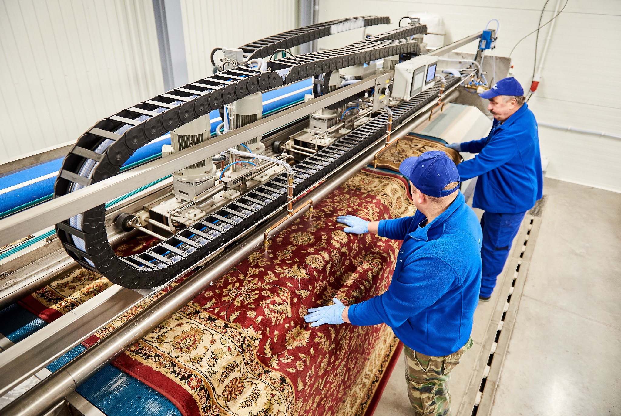 Dwóch mężczyzn w uniformach obsługuje automatyczną maszynę do prania dywanów z wzorem orientalnym w jasnym, przemysłowym pomieszczeniu.