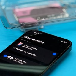 AppleHome - serwis iPhone Szczecin serwis Apple naprawa MacBook iPad - Smartfon iPhone 14 Pro z włączonym trybem diagnostycznym na tle niebieskiego podłoża i pojemnika z baterią. Widoczne pęknięcie na ekranie.