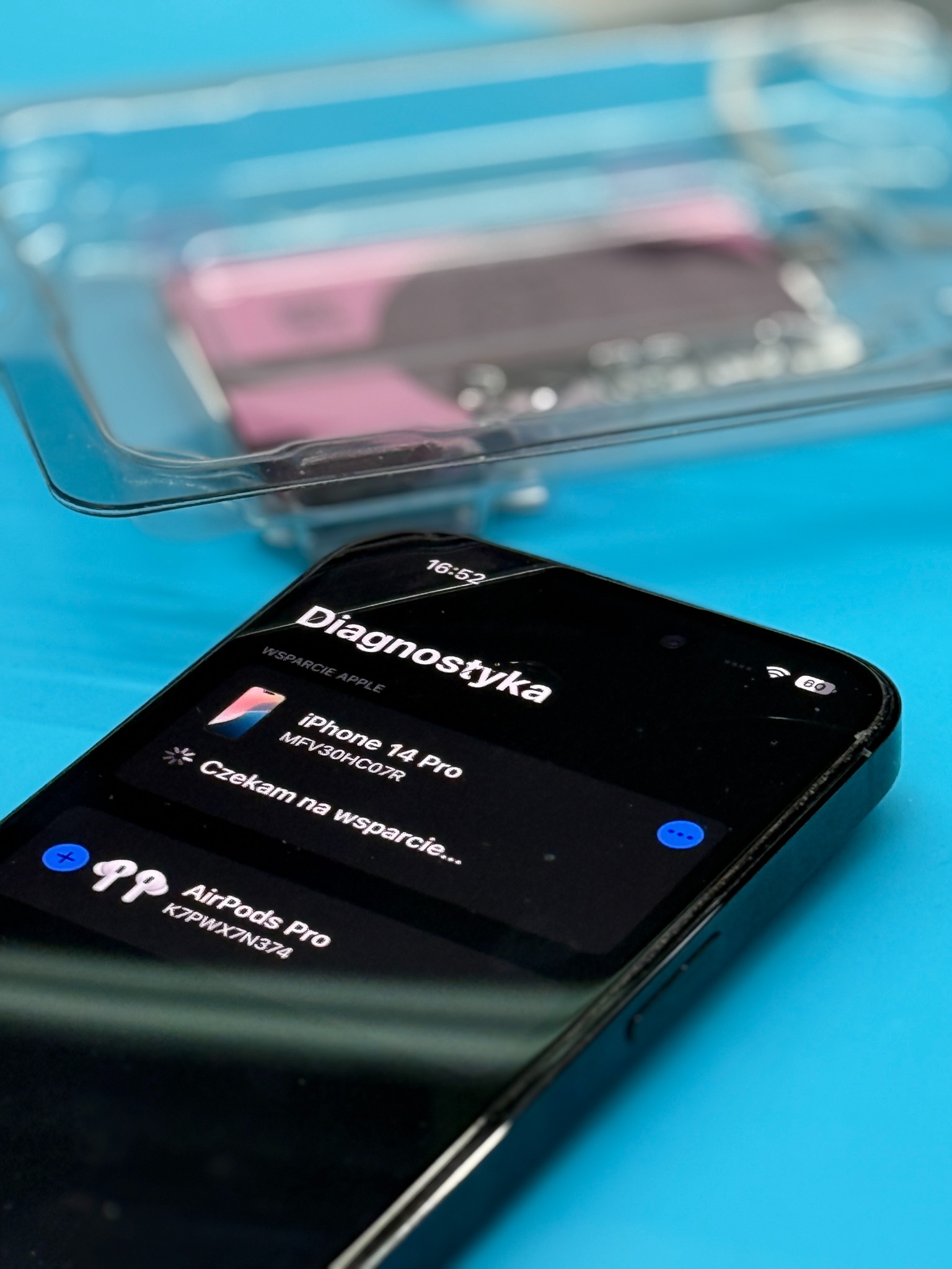 Smartfon iPhone 14 Pro z włączonym trybem diagnostycznym na tle niebieskiego podłoża i pojemnika z baterią. Widoczne pęknięcie na ekranie.