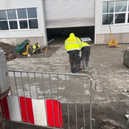 Układanie kostki brukowej na mokro. Pracownicy w odblaskowych ubraniach ubijają kostkę, w tle widoczny sprzęt budowlany i budynek z szarymi ścianami.