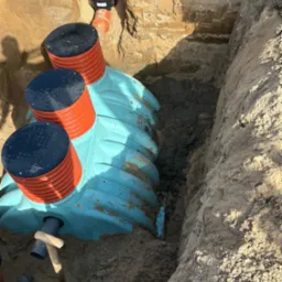 Montaż przydomowej oczyszczalni ścieków w wykopie. Widoczne elementy instalacji: zbiornik z pokrywami, rury kanalizacyjne, narzędzie do kopania.