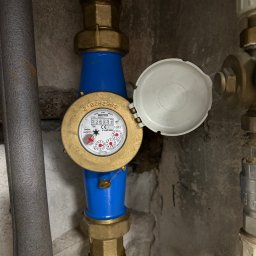 Zakład rzemieślniczy Popa & Kwiatkowski - Zamknięty licznik wody z niebieskimi rurami i mosiężnymi złączkami na tle szarej ściany. Widoczny fragment izolowanej rury i zaworu.