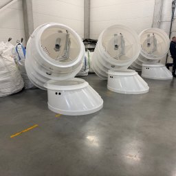 Białe, plastikowe elementy szamb betonowych w hali magazynowej, widoczne worki i dwie osoby w tle. Elementy ułożone na szarym betonowym podłożu.