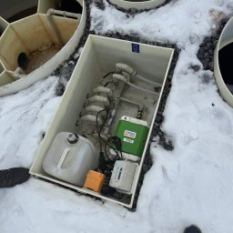 Otwarty zbiornik przydomowej oczyszczalni ścieków na śniegu, widoczne elementy instalacji, rury i urządzenia. Ujęcie z góry, fragment buta w kadrze.