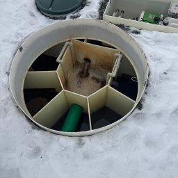 Otwarty zbiornik oczyszczalni ścieków na śniegu, widoczne elementy instalacji i pokrywa. Zielona rura wewnątrz zbiornika. Biała obudowa z elektroniką w tle.