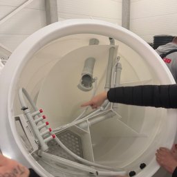 Wnętrze białego zbiornika z instalacją sanitarną, widoczne rury i elementy z tworzywa. Palec wskazuje na jeden z elementów. Widok z góry, oświetlenie hali.