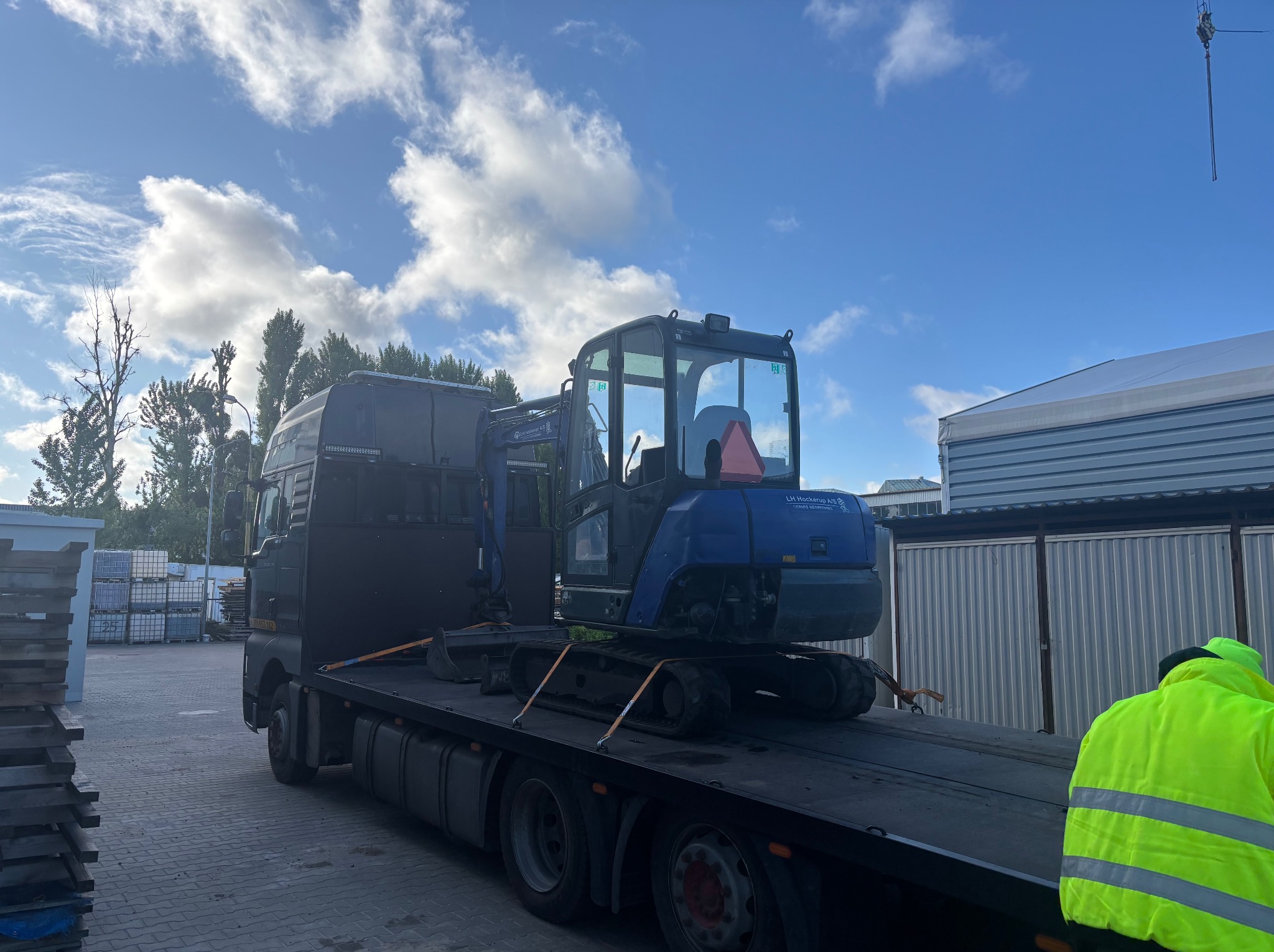 Niebieska mini koparka na lawecie ciężarówki, zabezpieczona pasami transportowymi, na tle błękitnego nieba z chmurami i budynków przemysłowych.