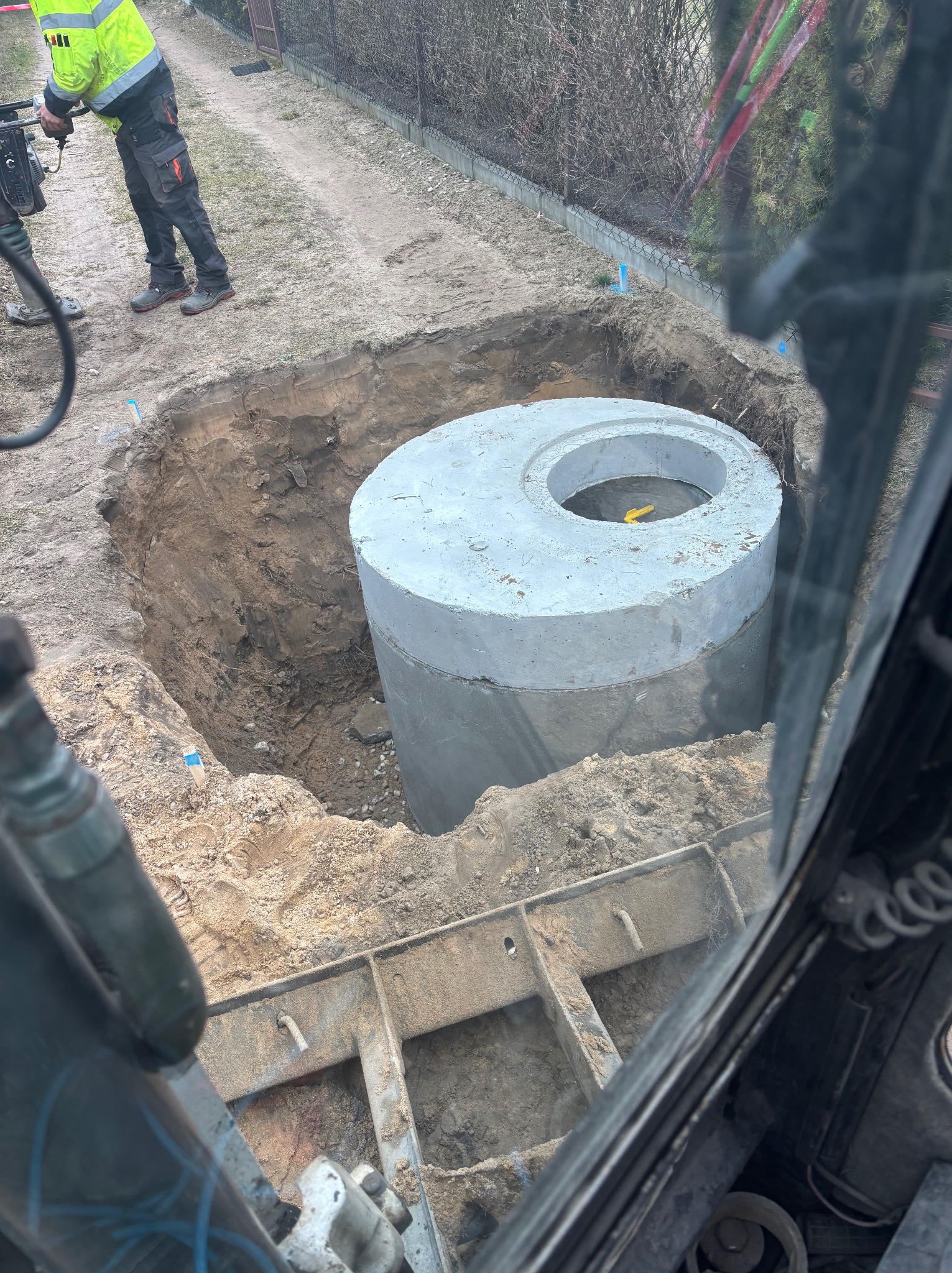 Betonowy zbiornik w wykopie, widok z wnętrza maszyny budowlanej. W tle ubijarka i fragment ogrodzenia. Etap montażu szamba.