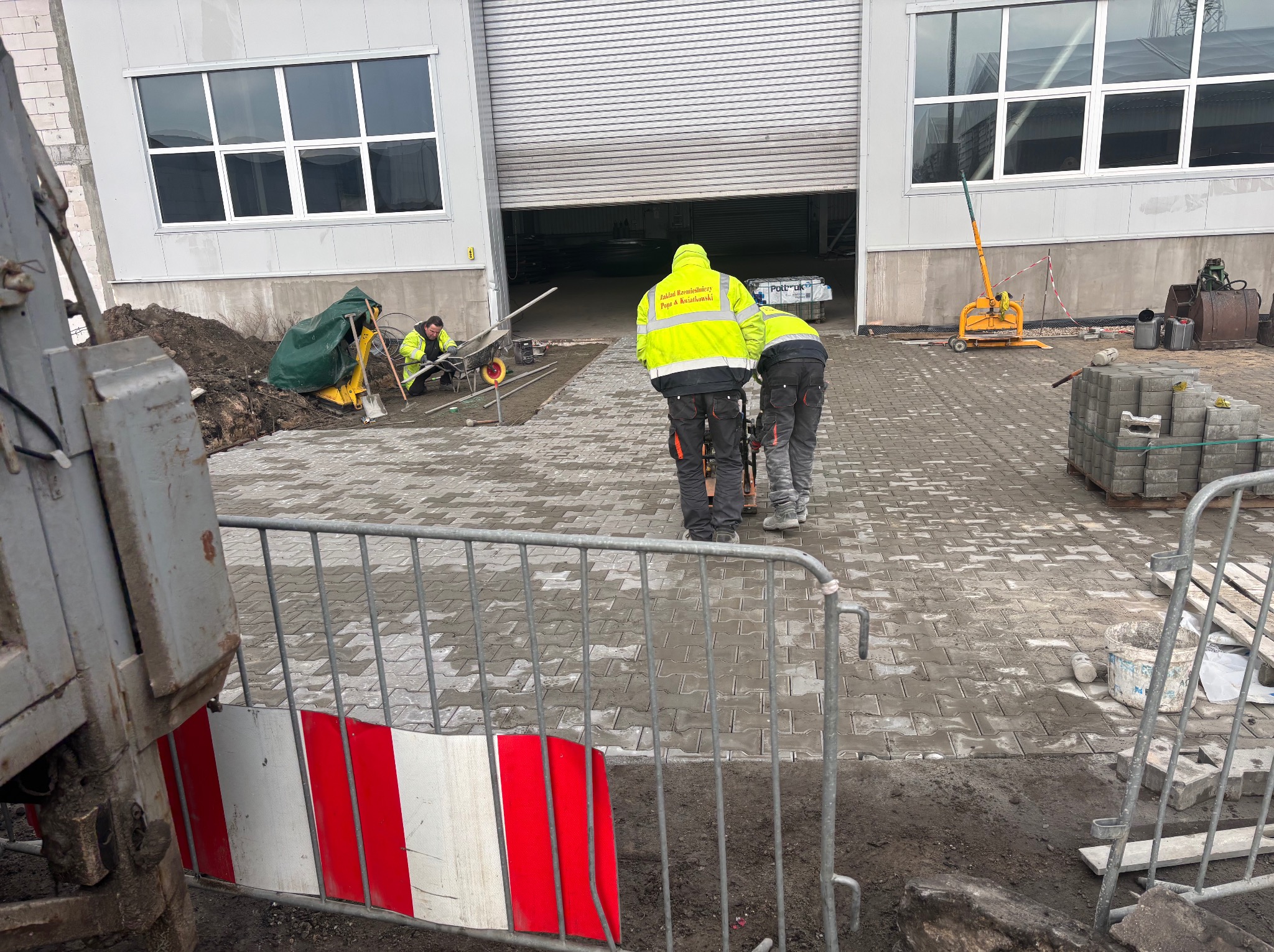 Układanie kostki brukowej na mokro. Pracownicy w odblaskowych ubraniach ubijają kostkę, w tle widoczny sprzęt budowlany i budynek z szarymi ścianami.