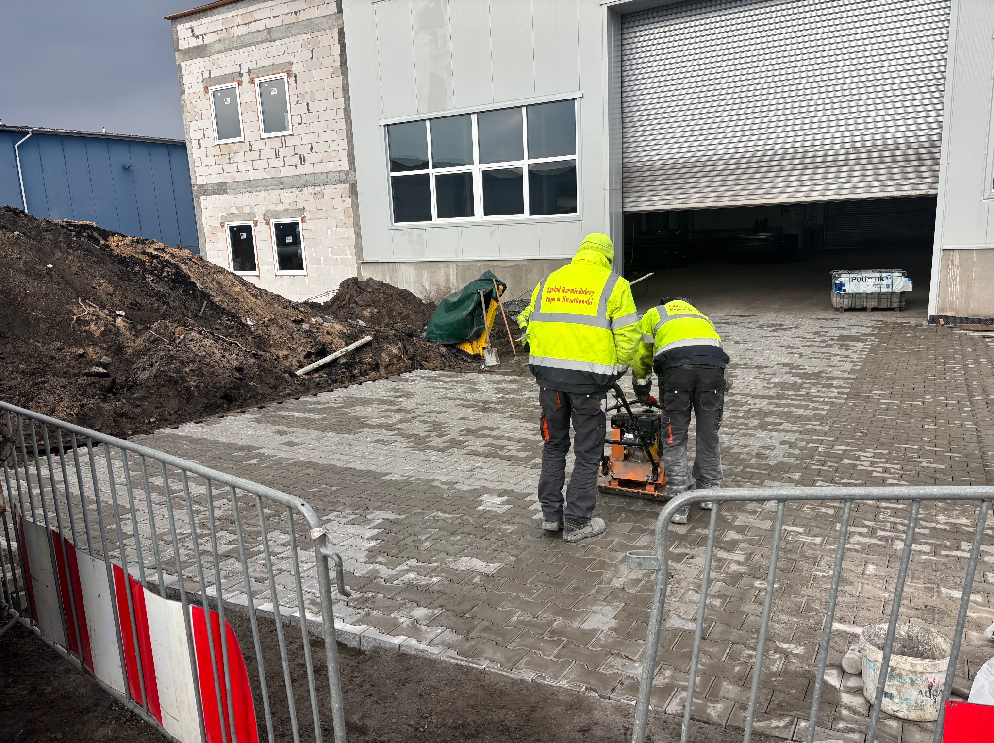 Pracownicy w odblaskowych ubraniach ubijają nowo ułożoną, szarą kostkę brukową zagęszczarką. W tle budynek przemysłowy i hałda ziemi.