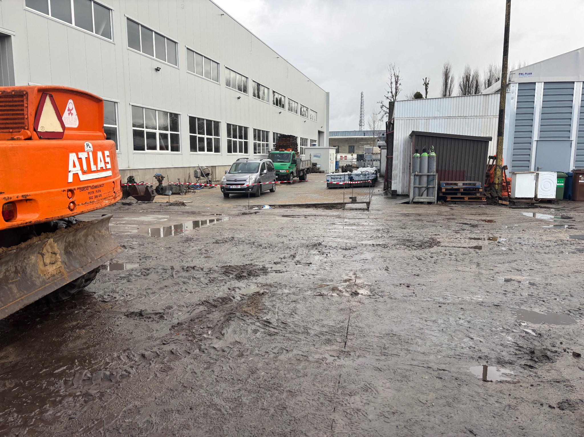 Pomarańczowa koparka Atlas na placu budowy z błotem, w tle budynek przemysłowy, samochody i materiały budowlane. Pochmurne niebo.