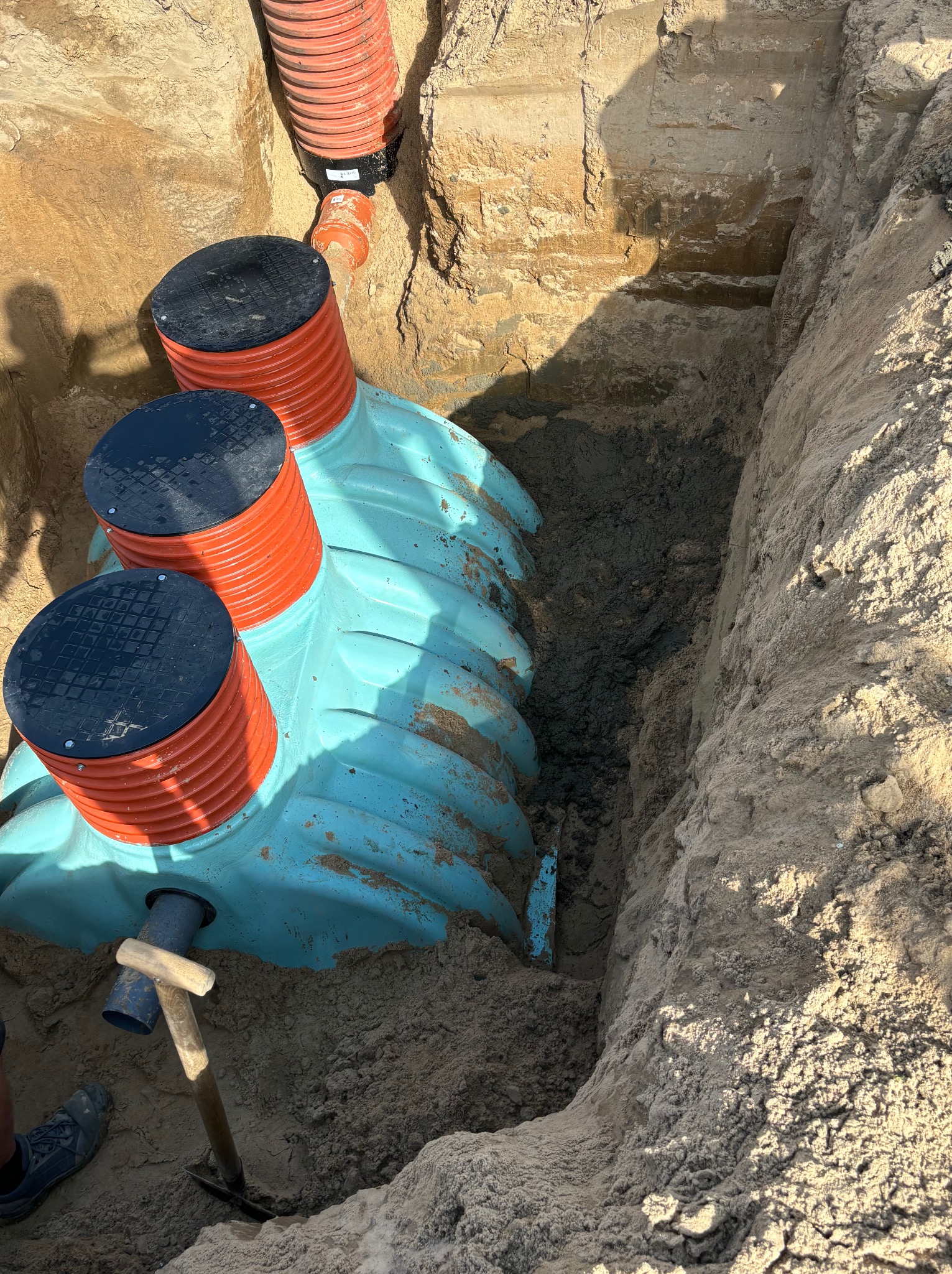 Montaż przydomowej oczyszczalni ścieków w wykopie. Widoczne elementy instalacji: zbiornik z pokrywami, rury kanalizacyjne, narzędzie do kopania.