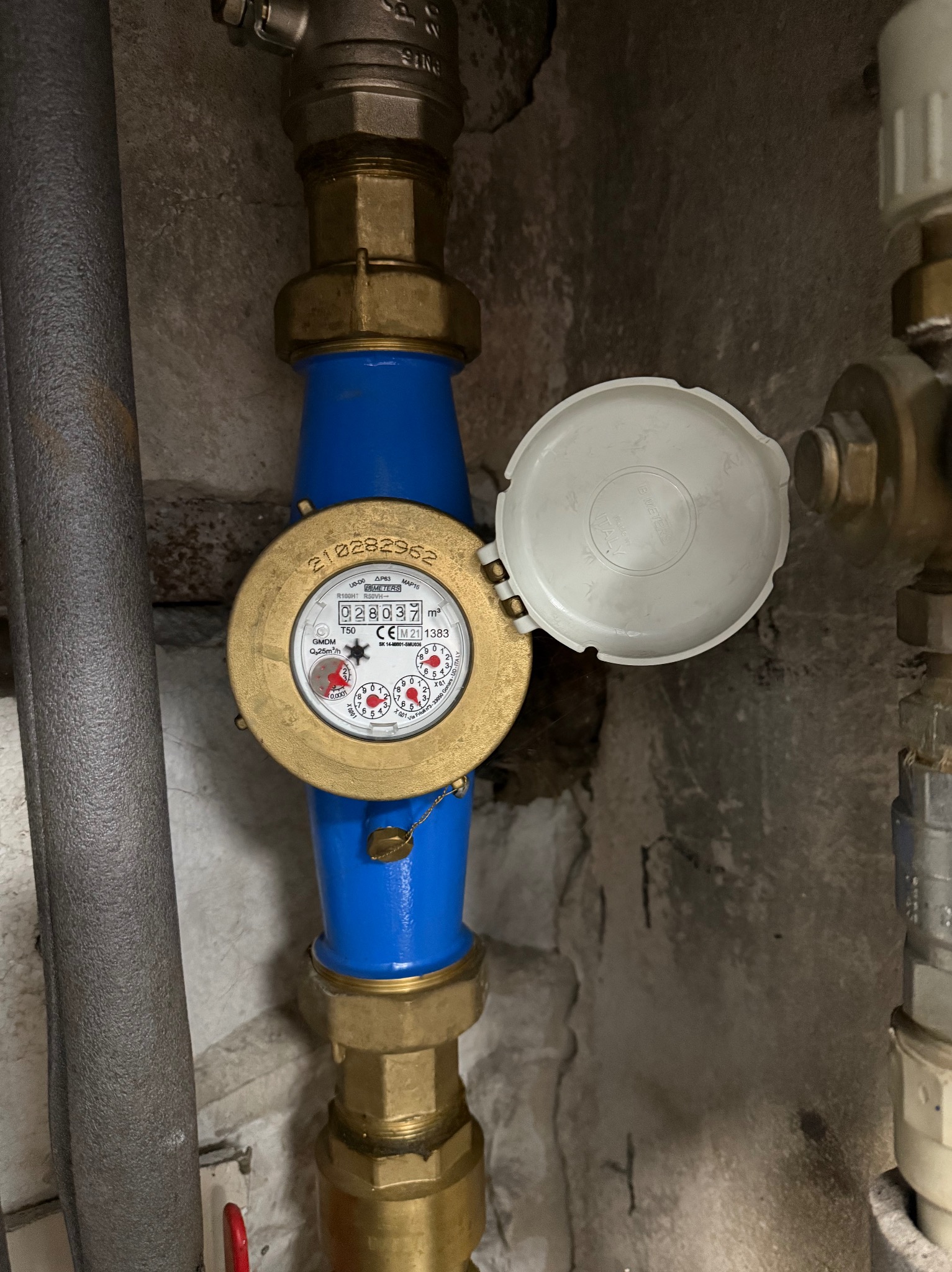 Zamknięty licznik wody z niebieskimi rurami i mosiężnymi złączkami na tle szarej ściany. Widoczny fragment izolowanej rury i zaworu.