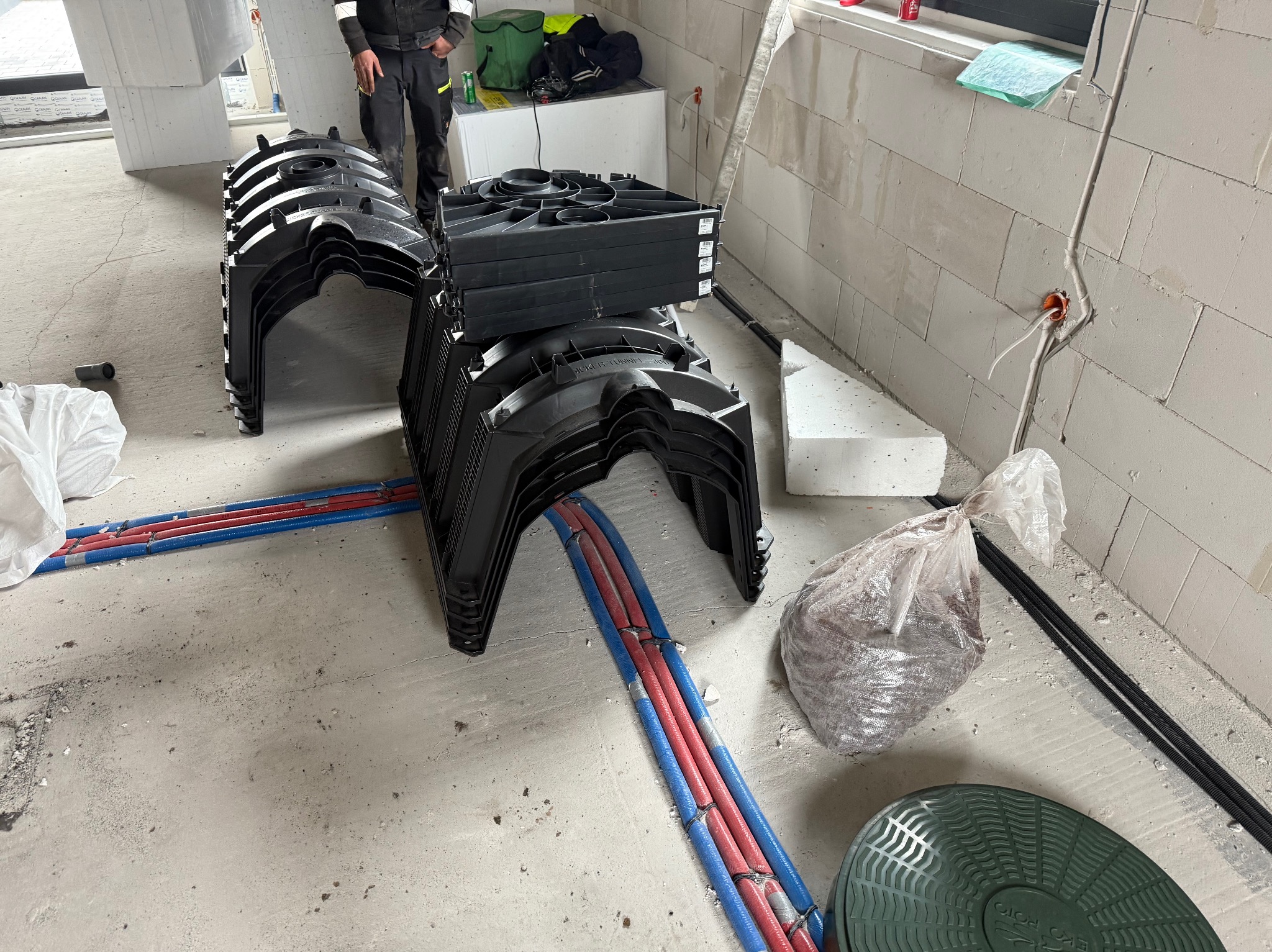 Montaż tuneli rozsączających Eko-Roto na budowie, widoczne rury instalacyjne i elementy konstrukcyjne budynku w surowym stanie.