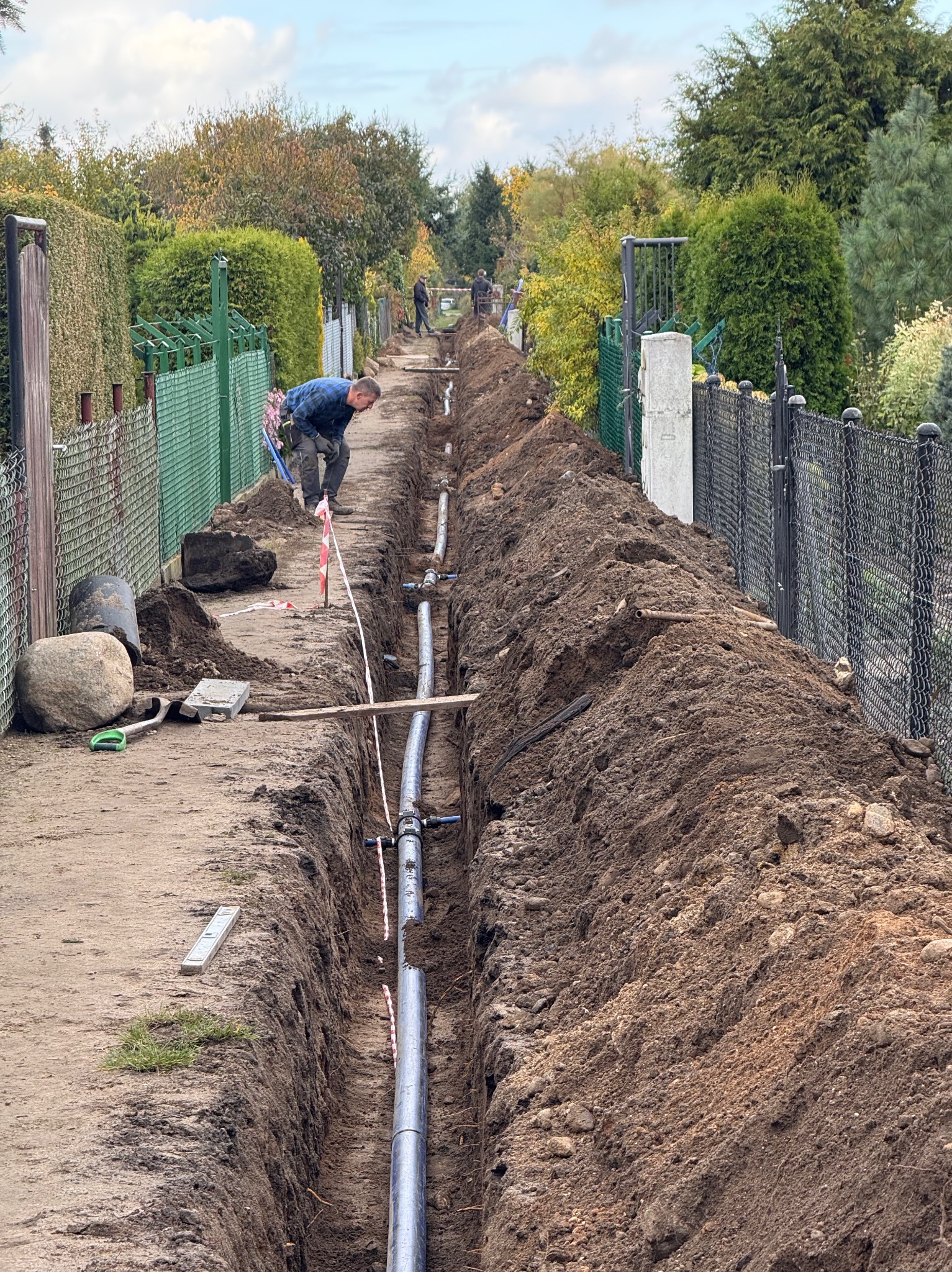 Wykopany rów z ułożonymi rurami, prawdopodobnie wodociągowymi, wzdłuż ogrodzenia posesji. Widoczny pracownik i narzędzia. Prace w toku.