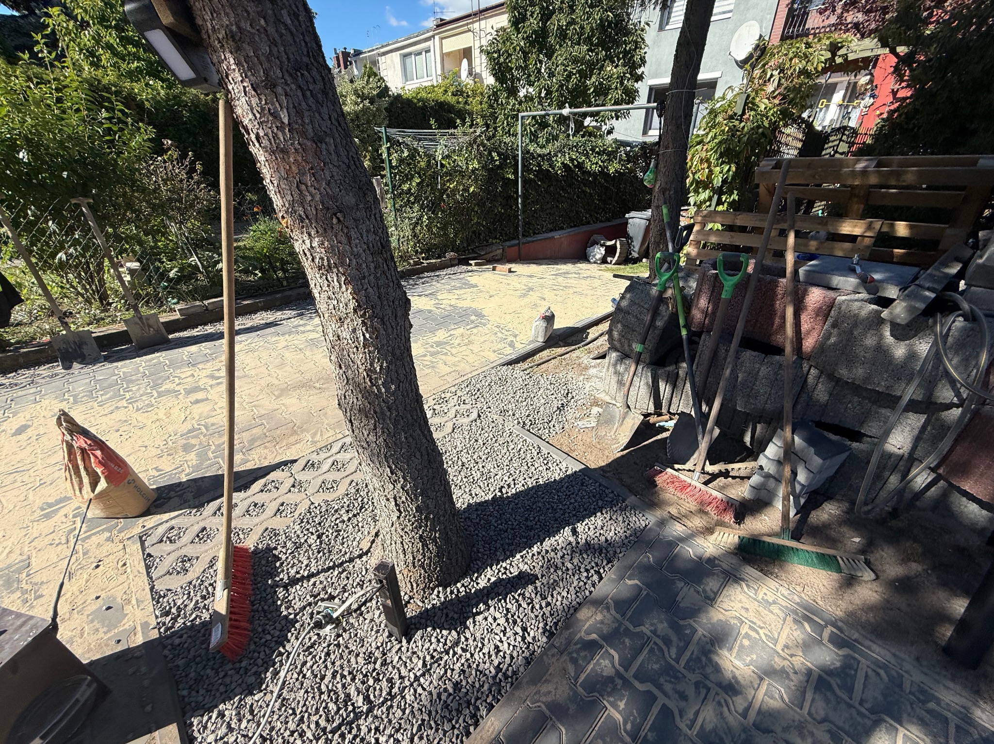 Układanie kostki brukowej wokół drzewa, widoczne narzędzia i materiały budowlane. Częściowo ułożona kostka, obok rozsypany żwir. Praca w toku, w tle dom i zieleń.