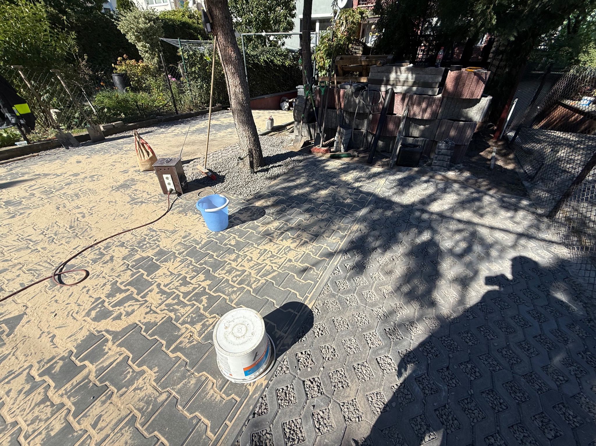 Układanie kostki brukowej: fragment ułożonej nawierzchni z kostki, częściowo zasypanej piaskiem, z widocznymi narzędziami i stosem kostki w tle.