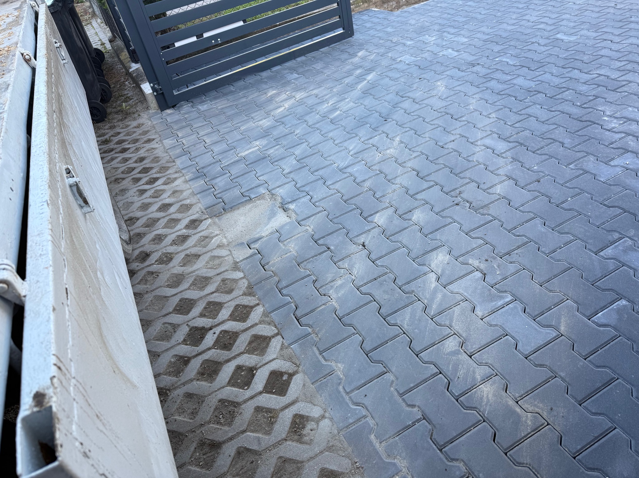 Ułożona szara kostka brukowa w jodełkę, częściowo na podłożu z ażurowych płyt. Fragment metalowej bramy w tle. Widoczne ślady użytkowania.