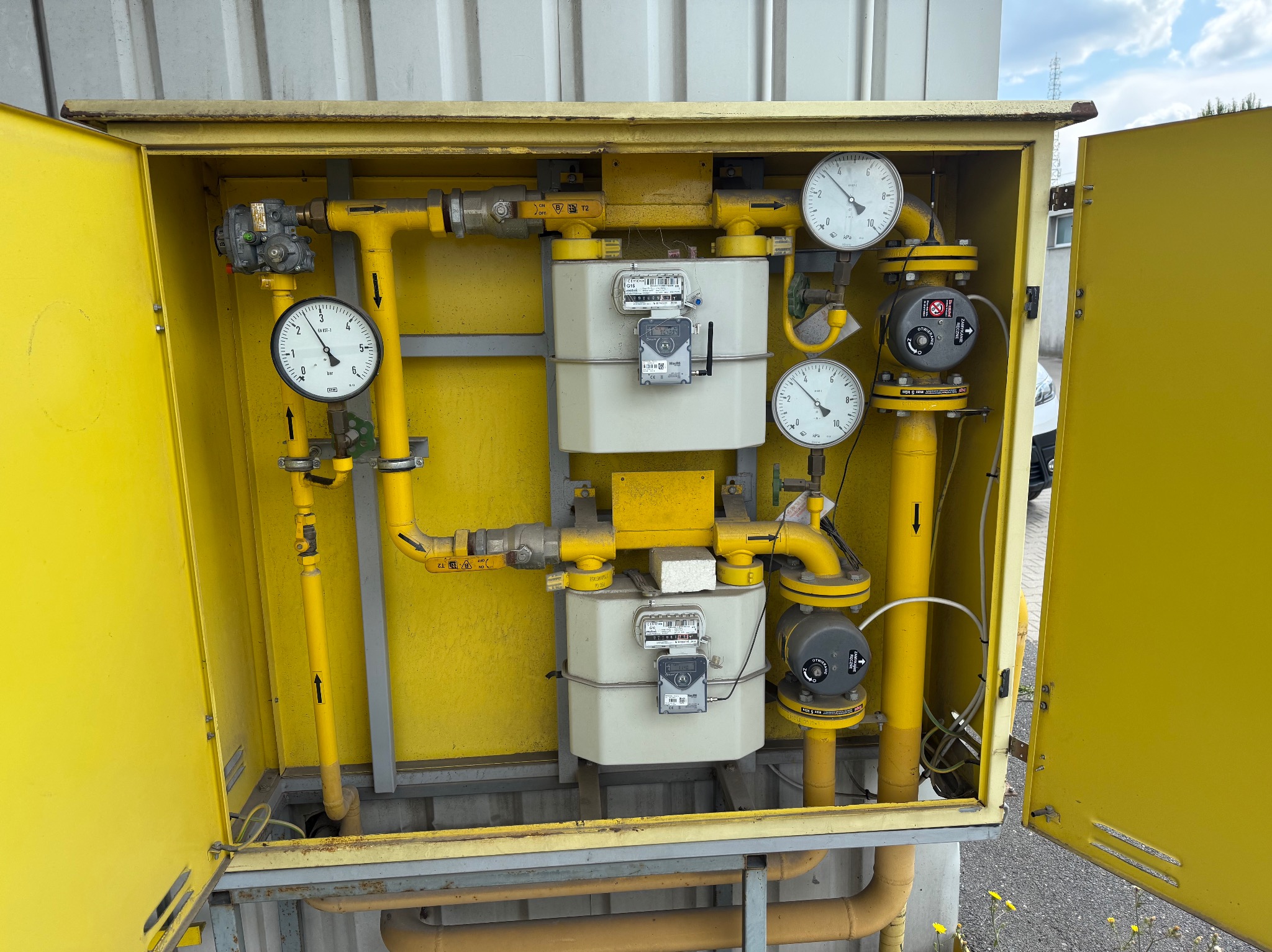 Żółta szafka gazowa z dwoma licznikami, manometrami i zaworami. Widoczne rury gazowe z oznaczeniami kierunku przepływu. Otwarta szafka, widoczne elementy montażowe.