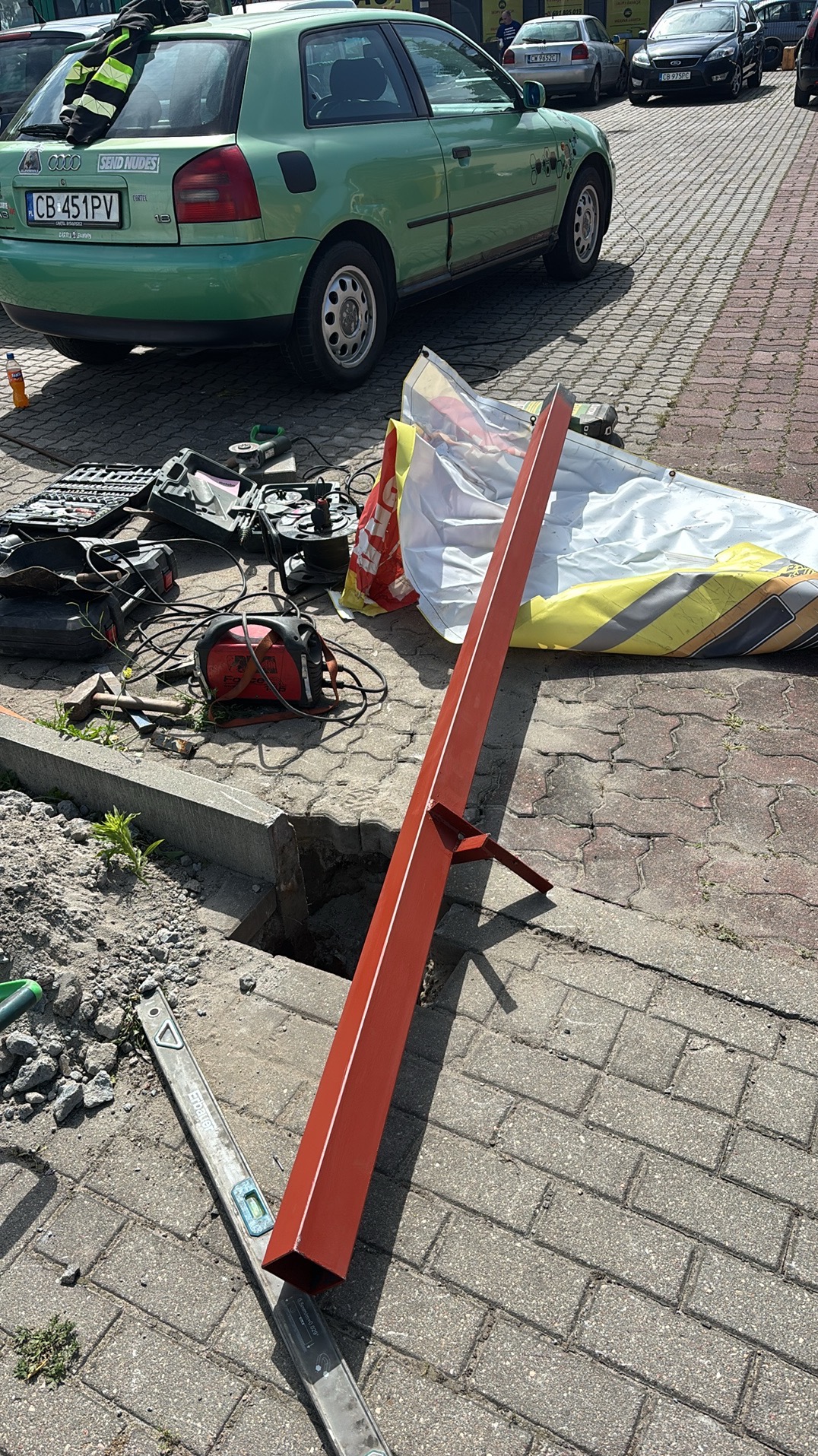 Wykop w kostce brukowej z narzędziami i profilem stalowym. Przygotowania do prac instalacyjnych na zewnątrz budynku. Widoczny poziomica i spawarka.