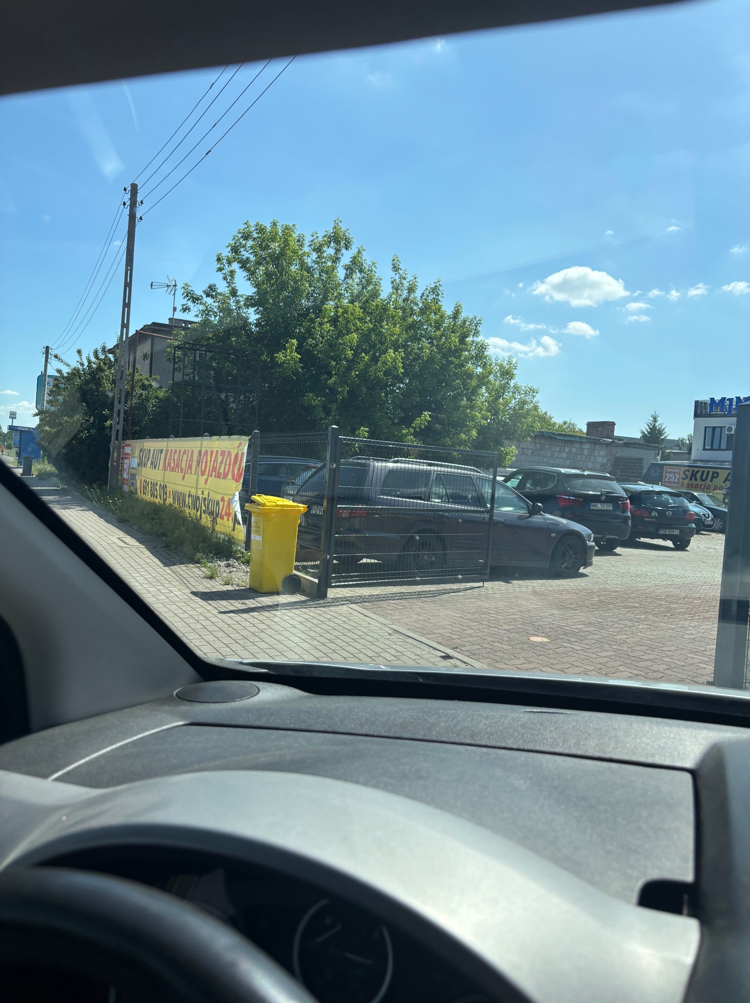 Widok z wnętrza samochodu na parking z autami i żółtym koszem na śmieci. W tle baner reklamowy skupu aut. Słoneczny dzień.