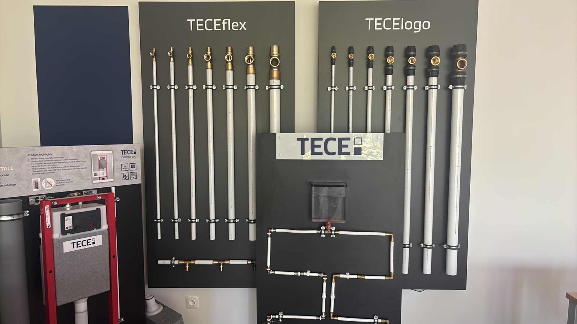 Ekspozycja systemów instalacyjnych TECEflex i TECElogo na ciemnoszarym tle. Prezentacja złączek, rur i modułu WC. Widoczne detale techniczne i elementy instalacyjne.