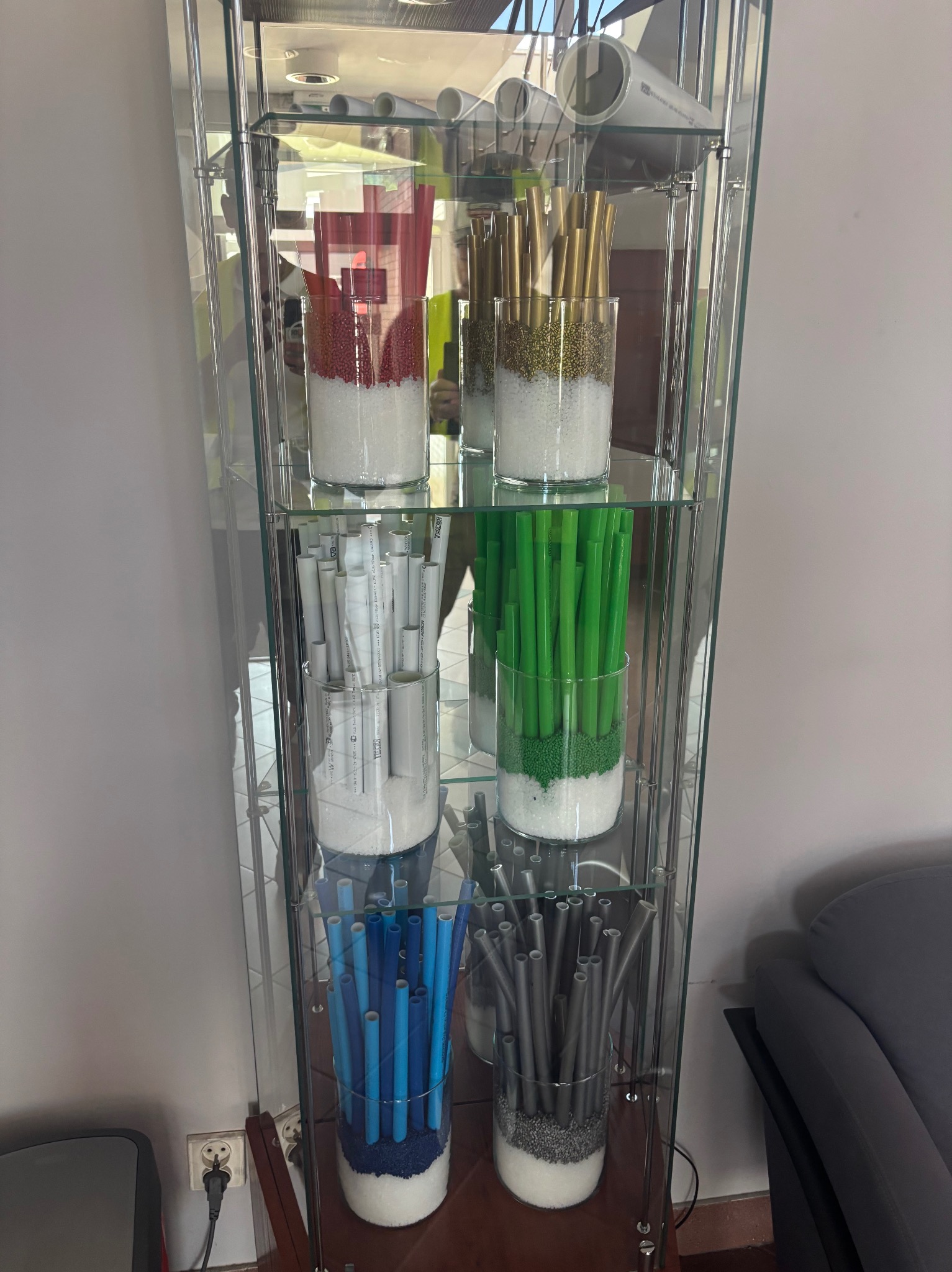 Szklana witryna z rurami PCV w różnych kolorach, umieszczonymi w szklanych wazonach wypełnionych białym i kolorowym granulatem. Dekoracja wnętrza z elementami instalacyjnymi.