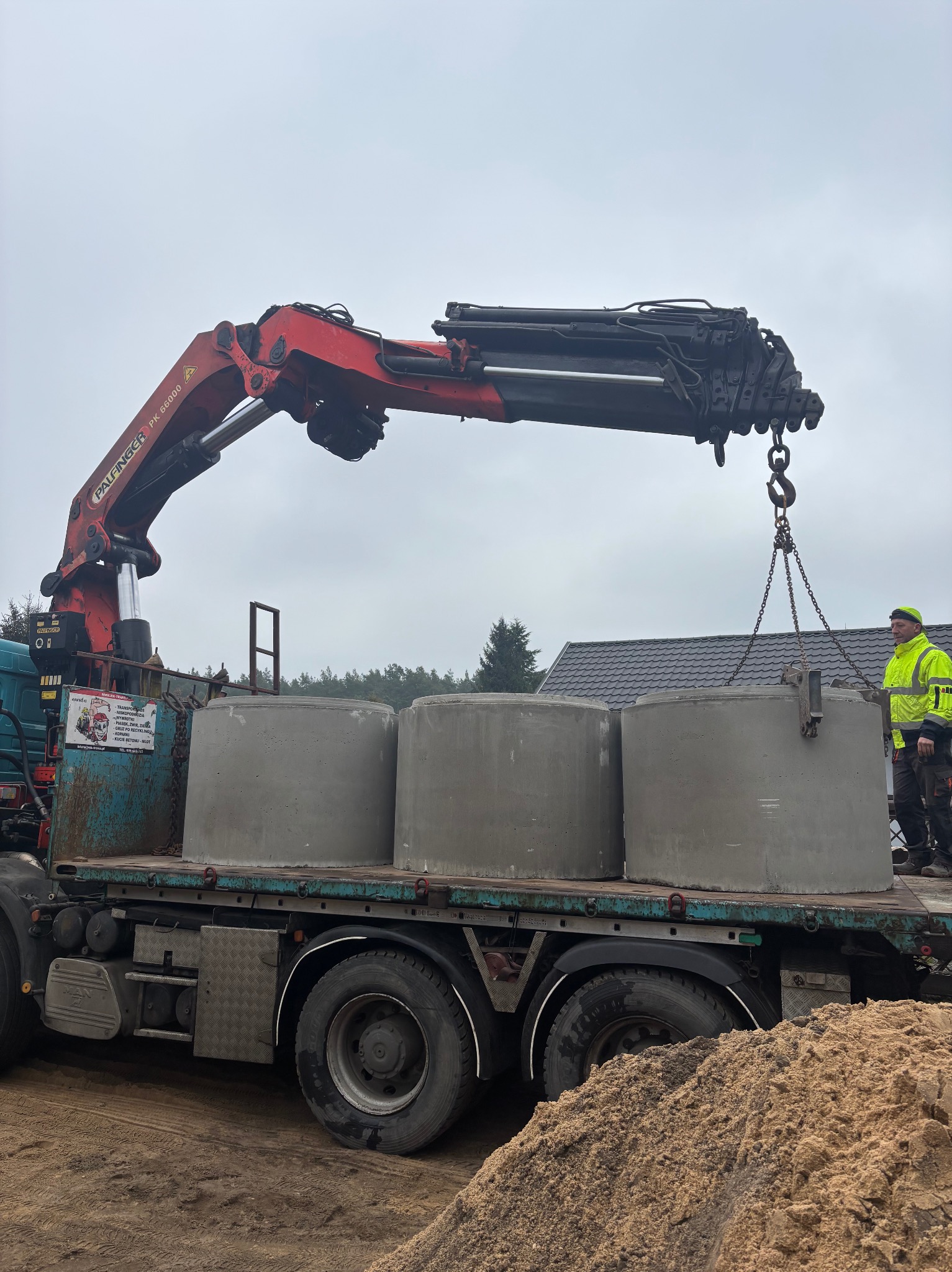 Transport szamba betonowego na ciężarówce z dźwigiem Palfinger PK 66000, pracownik w odblaskowej kurtce nadzoruje załadunek. W tle widoczny dom i drzewa.