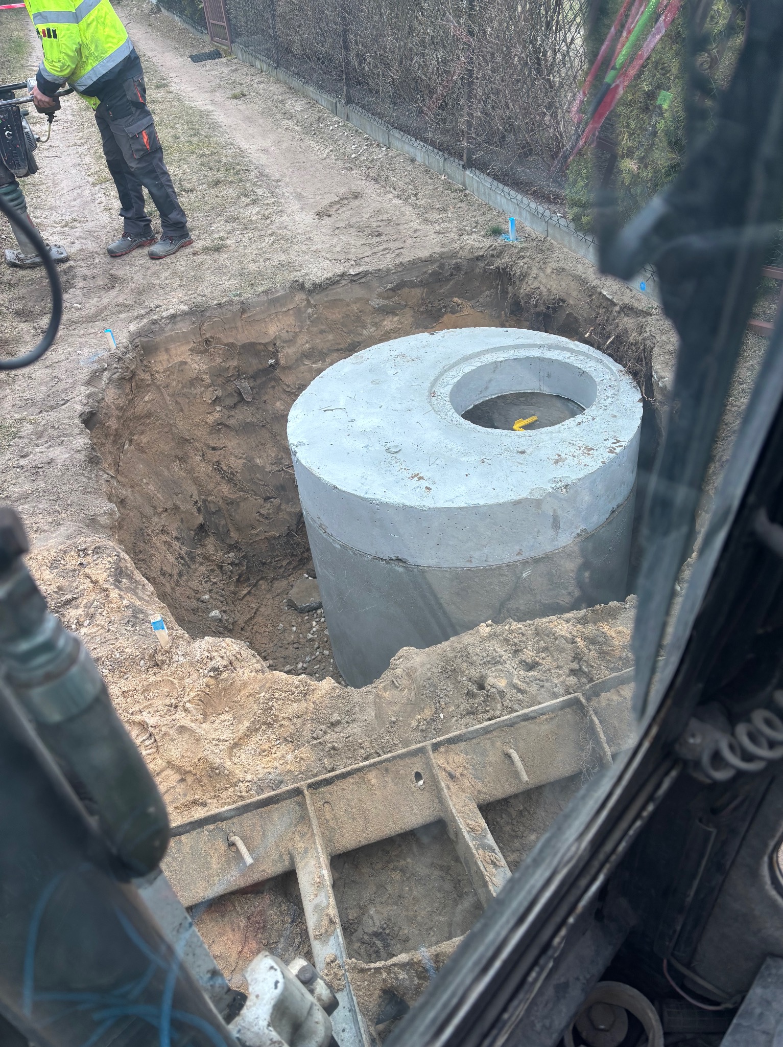 Betonowy zbiornik w wykopie, widok z kabiny maszyny budowlanej. W tle ubijarka i pracownik w odblaskowej kurtce. Prace ziemne w toku.