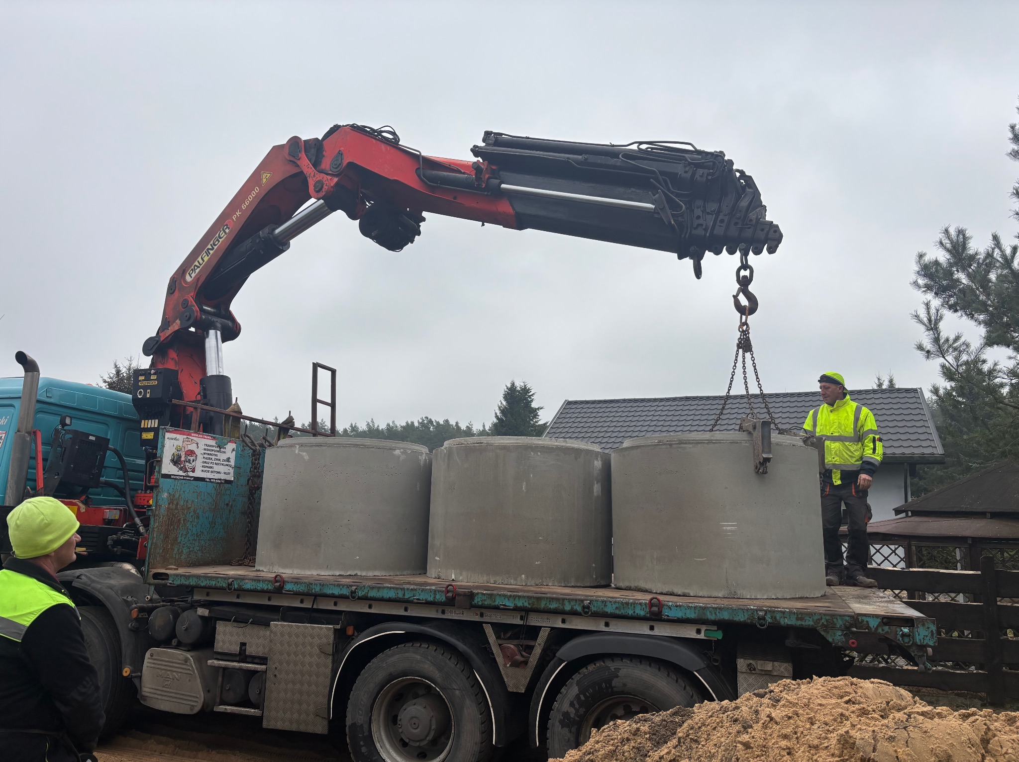 Transport szamba betonowego na ciężarówce z dźwigiem Palfinger PK 66000 A. Pracownik w odblaskowej kurtce nadzoruje załadunek. W tle dom i drzewa.