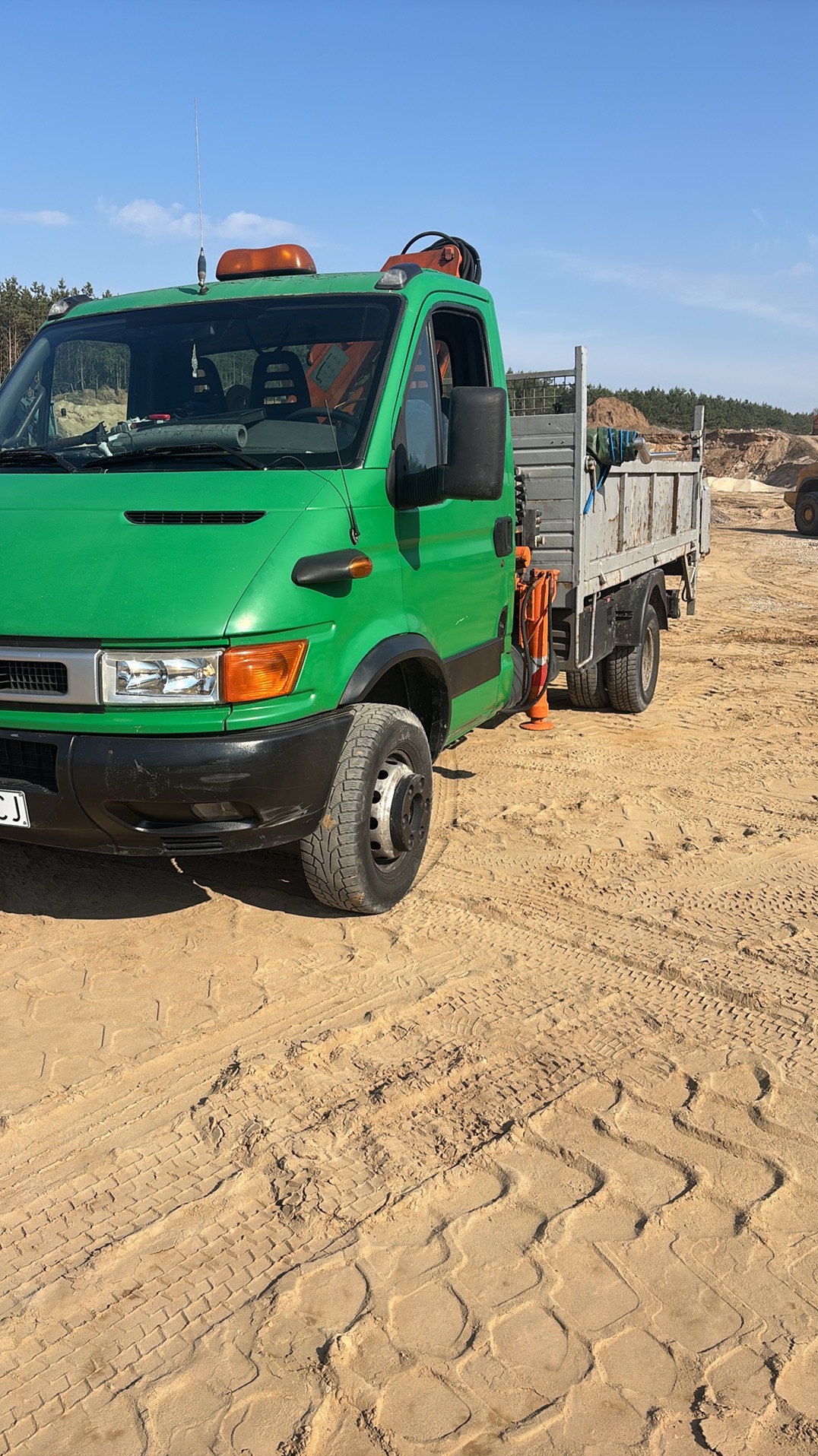 Zielona ciężarówka na piaszczystym terenie budowy, z widocznymi śladami opon i elementami krajobrazu w tle. Na pace leżą narzędzia.