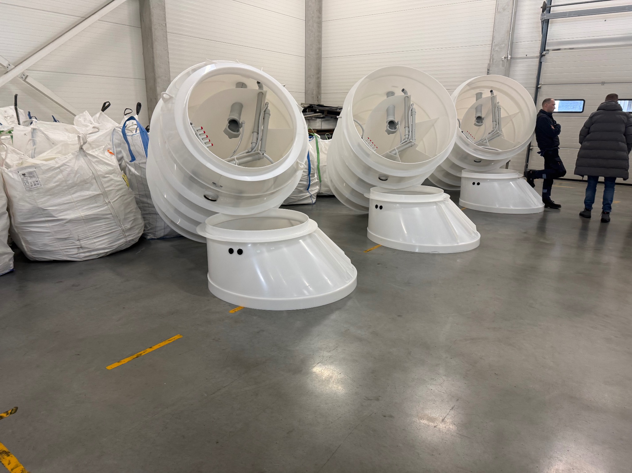 Białe, plastikowe elementy szamb betonowych w hali magazynowej, widoczne worki i dwie osoby w tle. Elementy ułożone na szarym betonowym podłożu.