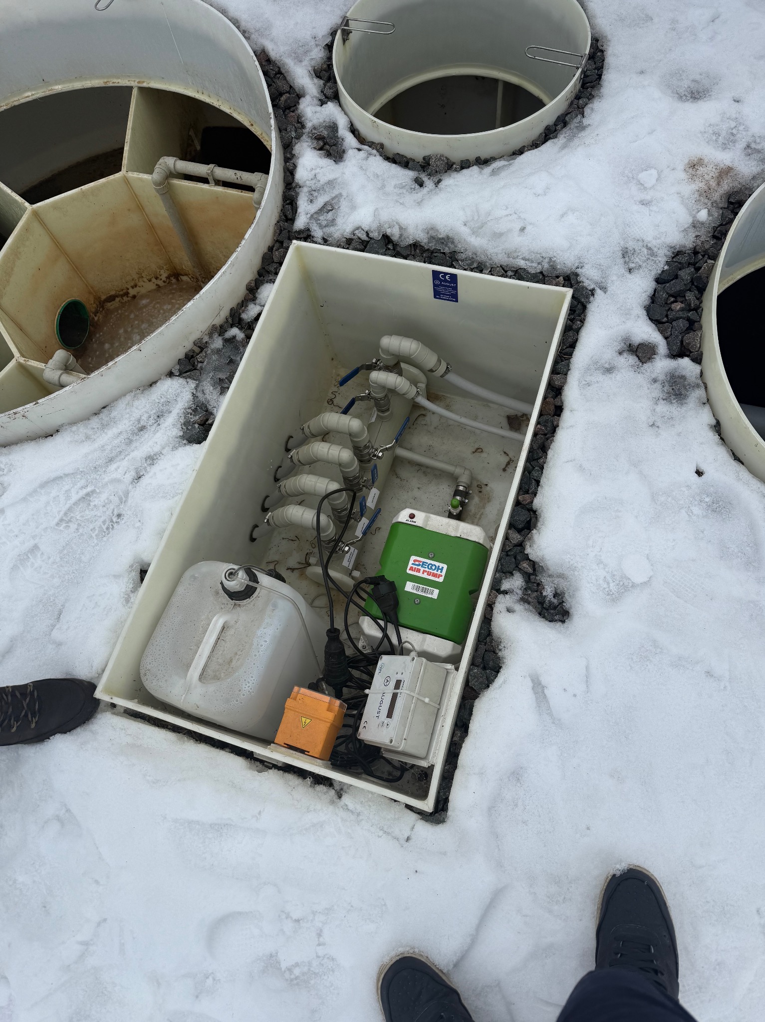 Otwarty zbiornik przydomowej oczyszczalni ścieków na śniegu, widoczne elementy instalacji, rury i urządzenia. Ujęcie z góry, fragment buta w kadrze.