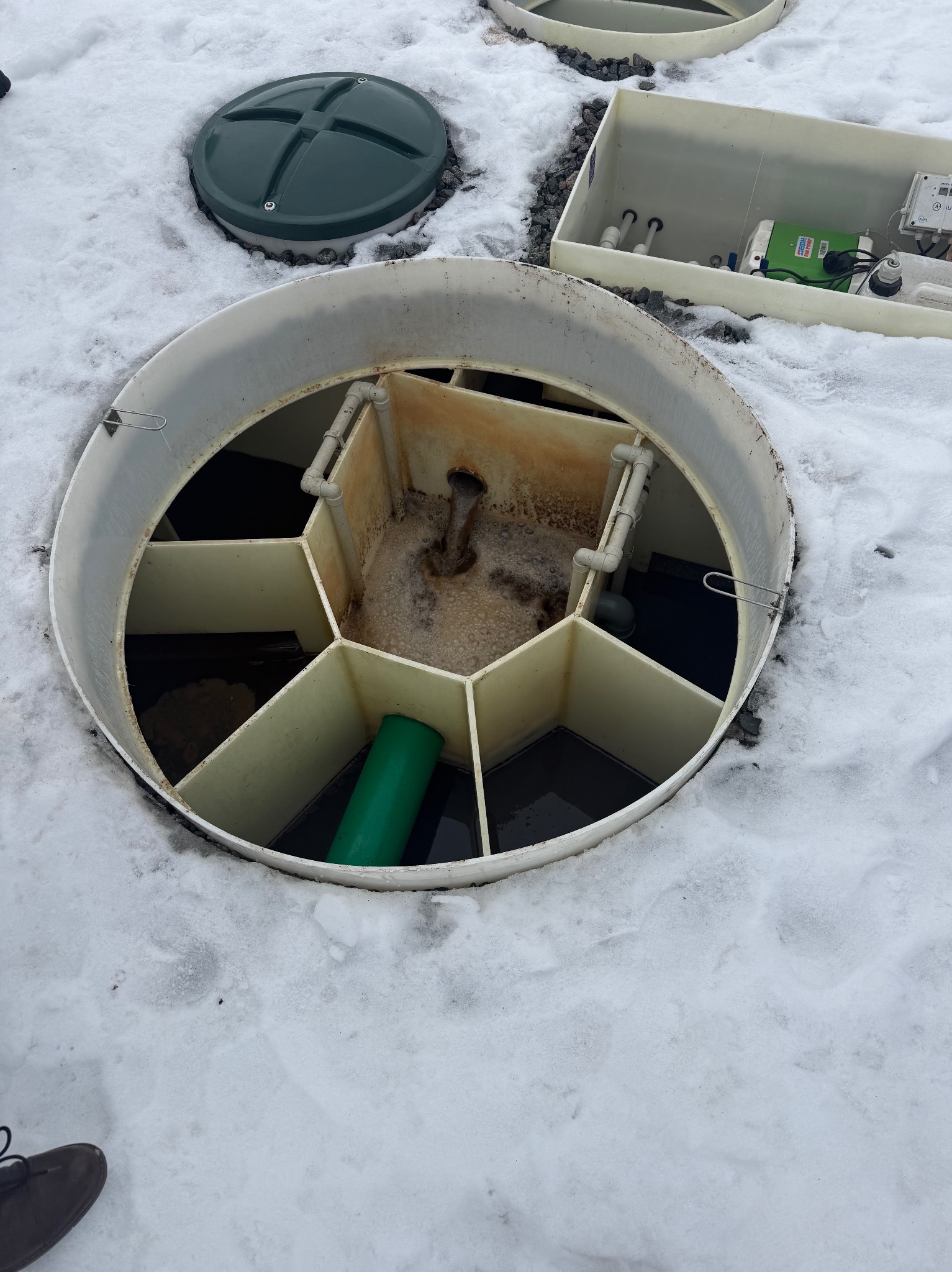 Otwarty zbiornik oczyszczalni ścieków na śniegu, widoczne elementy instalacji i pokrywa. Zielona rura wewnątrz zbiornika. Biała obudowa z elektroniką w tle.