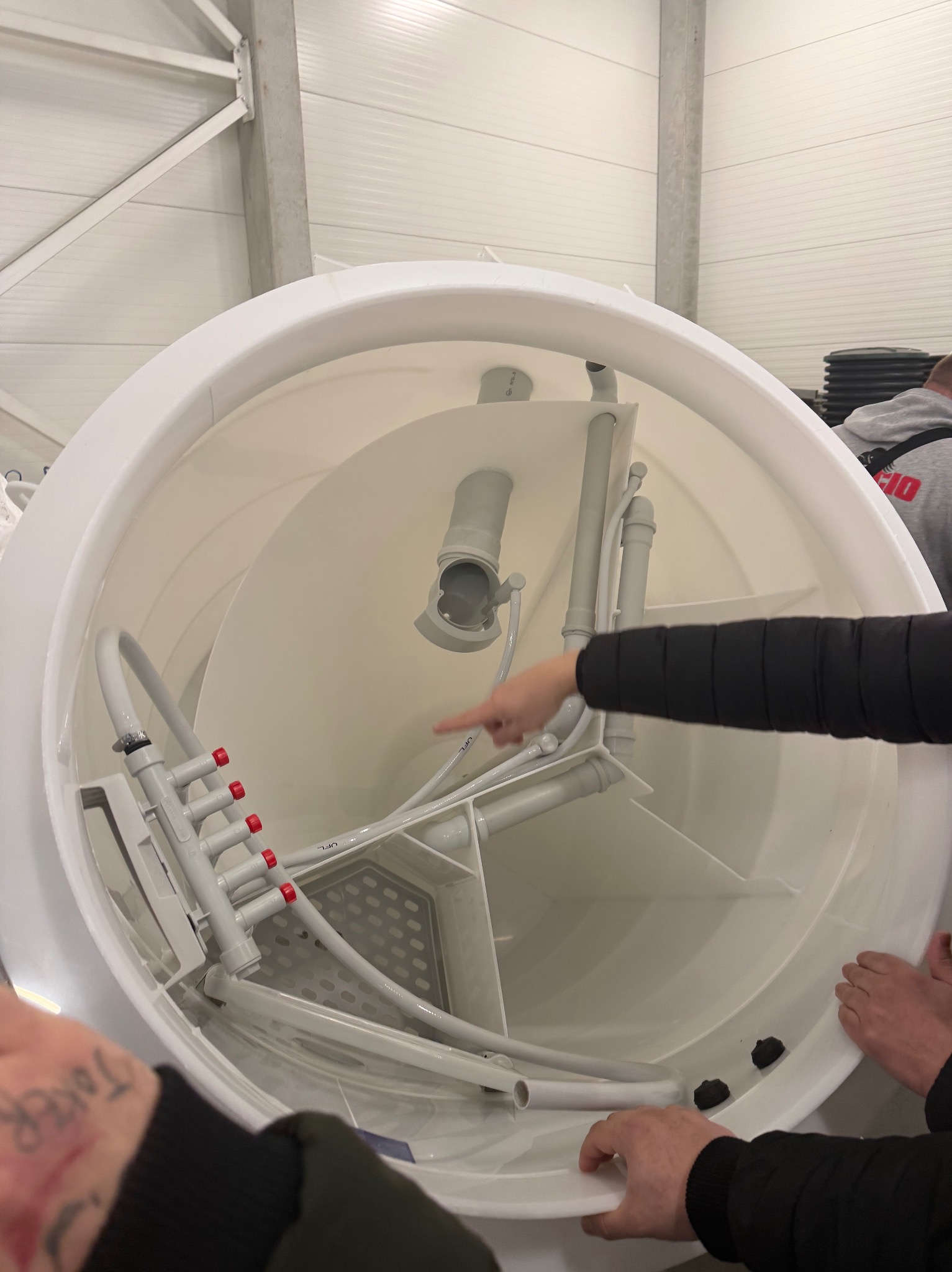 Wnętrze białego zbiornika z instalacją sanitarną, widoczne rury i elementy z tworzywa. Palec wskazuje na jeden z elementów. Widok z góry, oświetlenie hali.